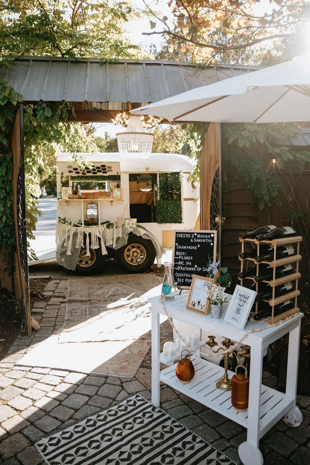 A vintage white trailer decorated for an outdoor event, with a menu board, wine bottles on a rack, and various decorative items. A white table with candles, flowers, and framed signs is in front, all set on a cobblestone pathway with a patterned rug. Sunlight filters through trees above.