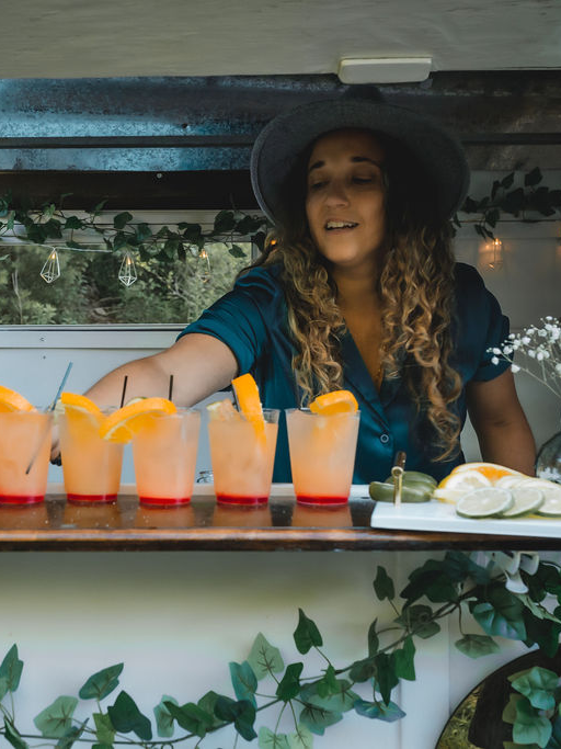 Woman preparing cocktails at outdoor bar with citrus garnishes and greenery.