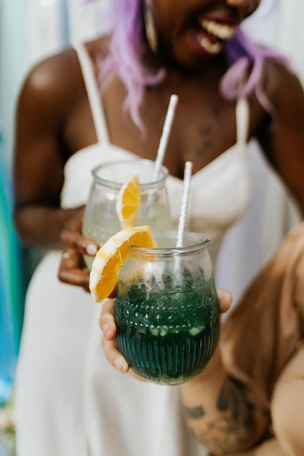 A woman with purple hair holding a green beverage garnished with an orange slice, smiling and wearing a white top.