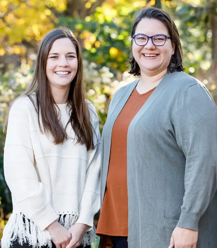 Two women standing outdoors in a park, smiling at the camera with trees and sunlight in the background.