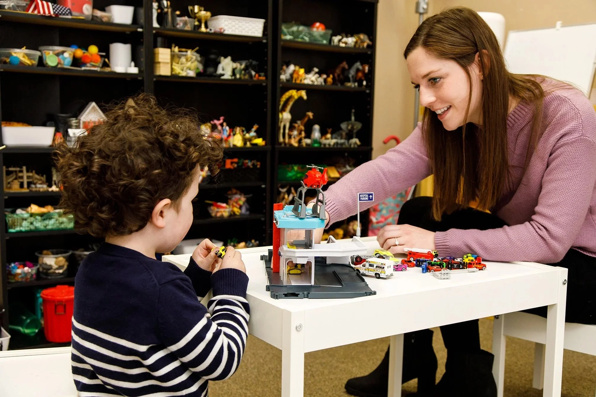 A woman and a young boy playing with toy cars at a white table in a toy or collectibles store. The woman is smiling and leaning over the table, while the boy is focused on the cars. Behind them are black shelves filled with various toys and collectibles.