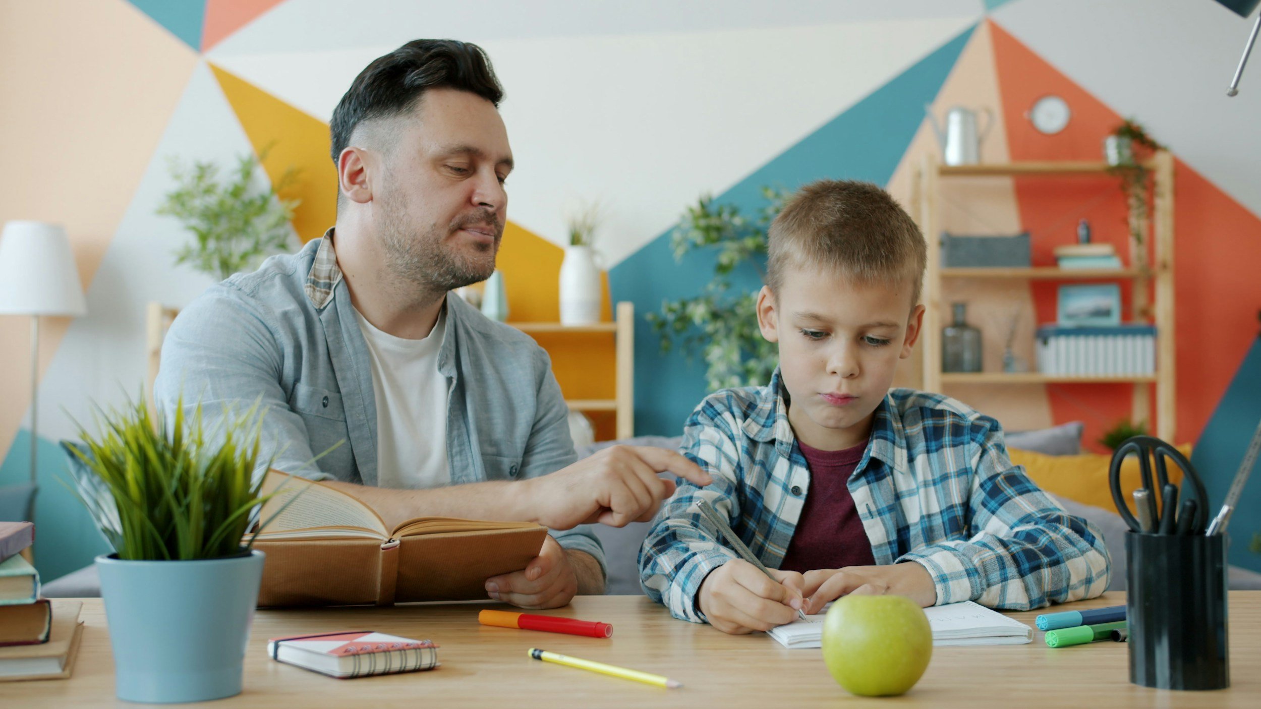 Image of father helping son with homework. The son looks focused but bored. The father is peering over and looking at the son's work.