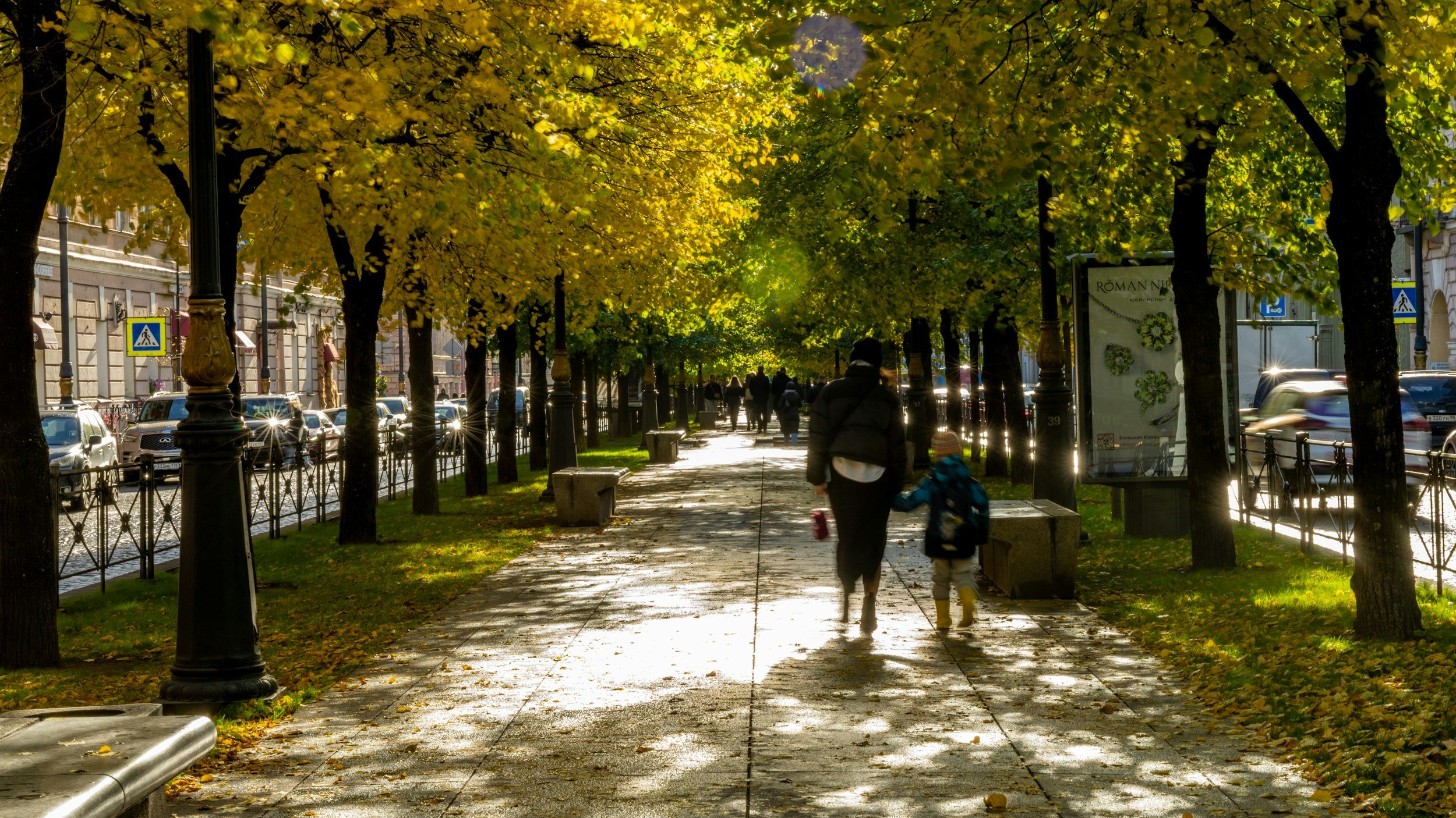Photo of woman and child holding hands walking down urban path surrounded by autumn trees.