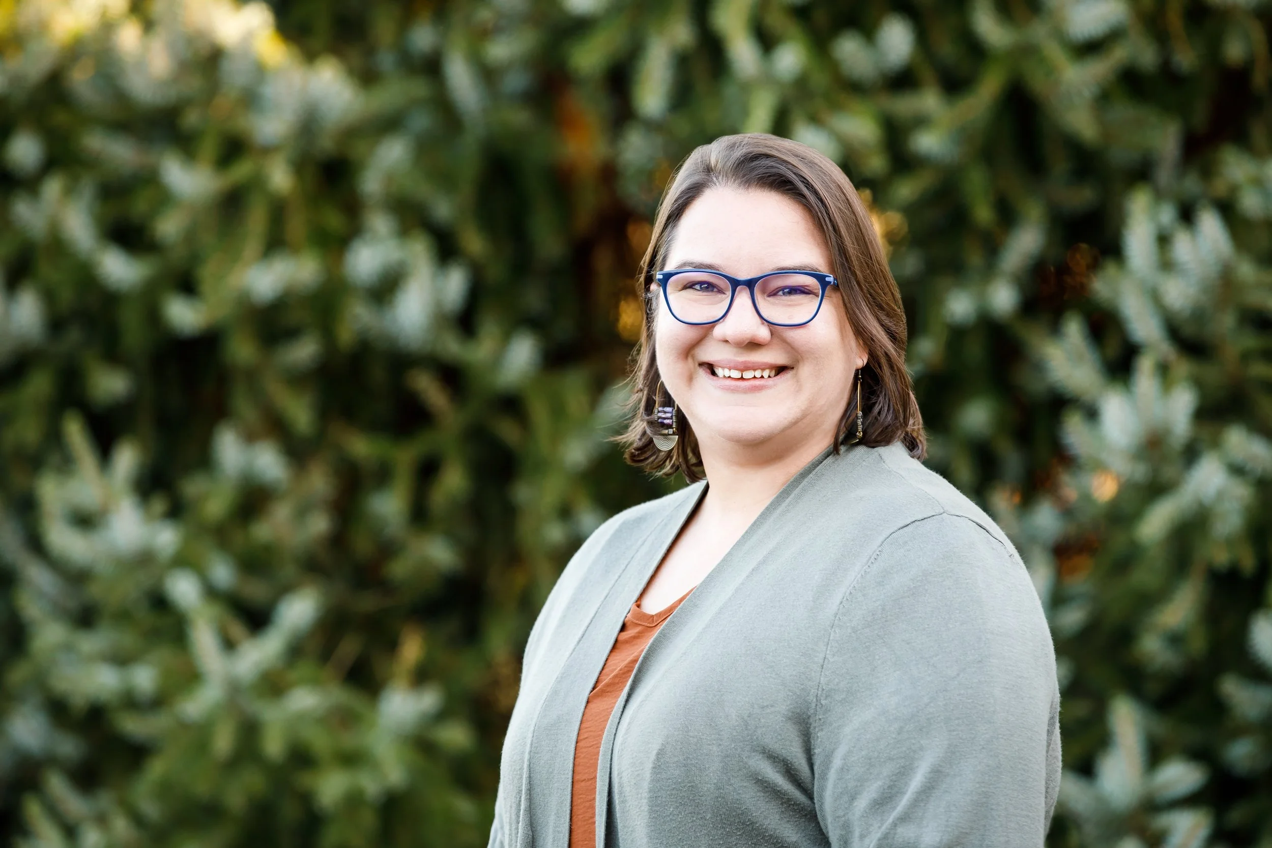 Photo of Jenny Liu, LPCC-S standing in front of evergreen trees. Jenny Liu is the owner of Conscious Roots Counseling in Blue Ash Ohio. She wears glasses and is smiling.
