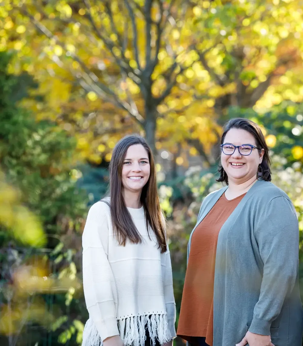 Two women smiling outdoors with a tree with yellow and green leaves in the background.