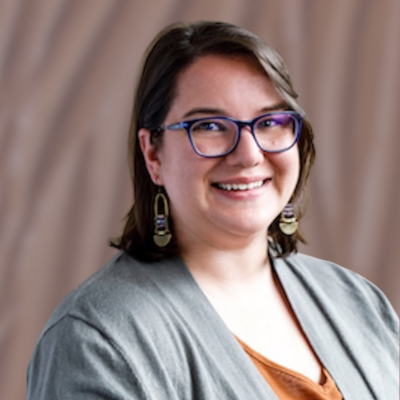 Jenny Liu, LPCC-S is a therapist and owner of Conscious Roots Counseling. She looks like a smiling woman with glasses and earrings, wearing a gray jacket and an orange top, with a blurred beige curtain background.