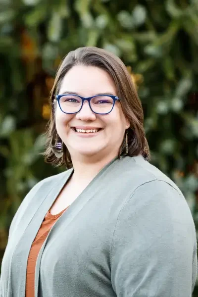A woman with shoulder-length brown hair, glasses, and earrings smiling outdoors with green foliage in the background.