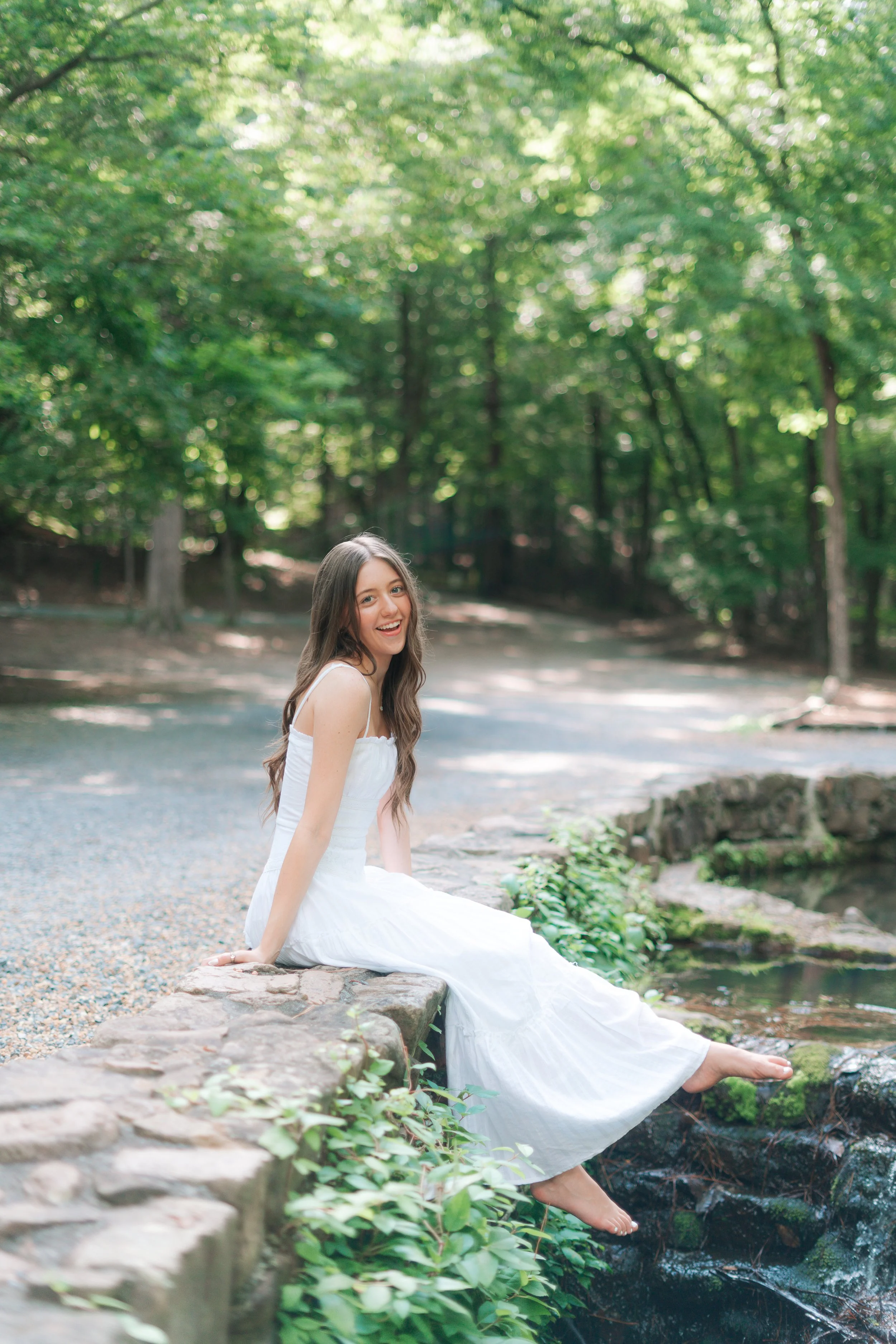 A young woman in a white dress sitting on a stone ledge by a small stream in a lush green forest, smiling at the camera.