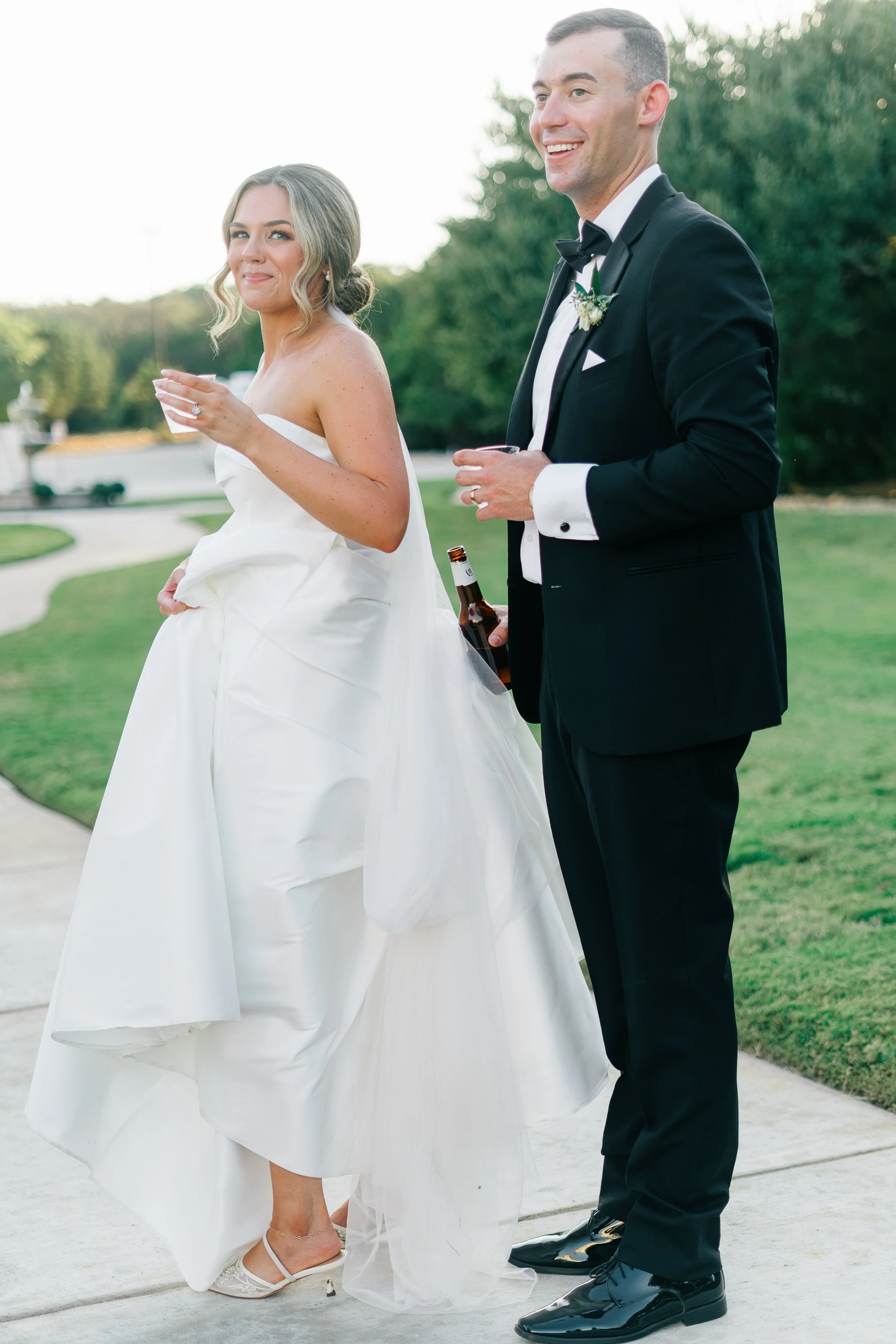 A bride and groom stand outdoors during their wedding reception, holding drinks and smiling, with trees and a paved walkway in the background.