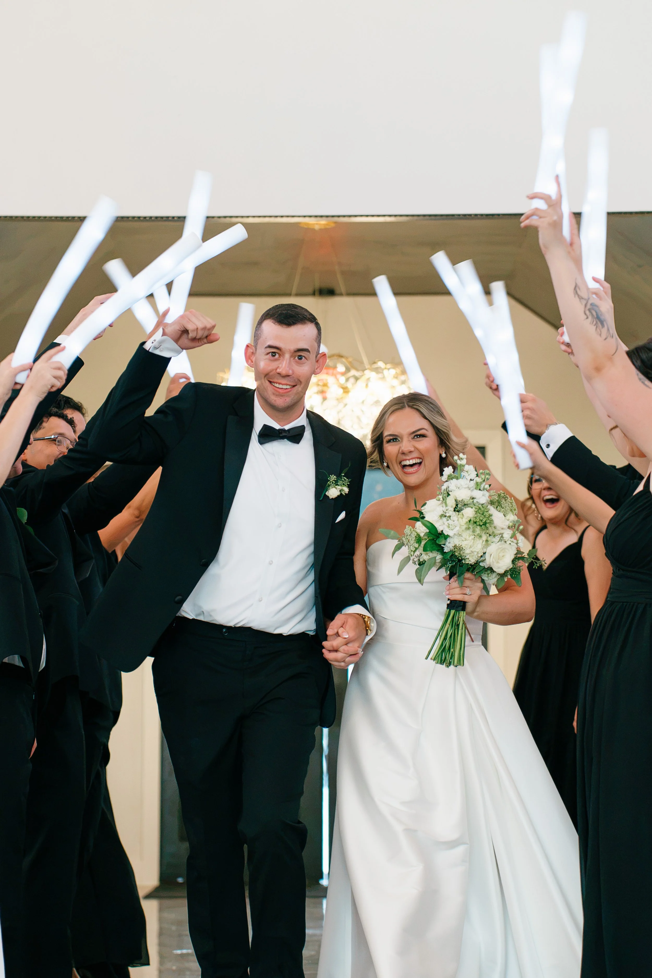 A newlywed couple holding hands and smiling, surrounded by friends celebrating indoors, with some holding illuminated sticks.