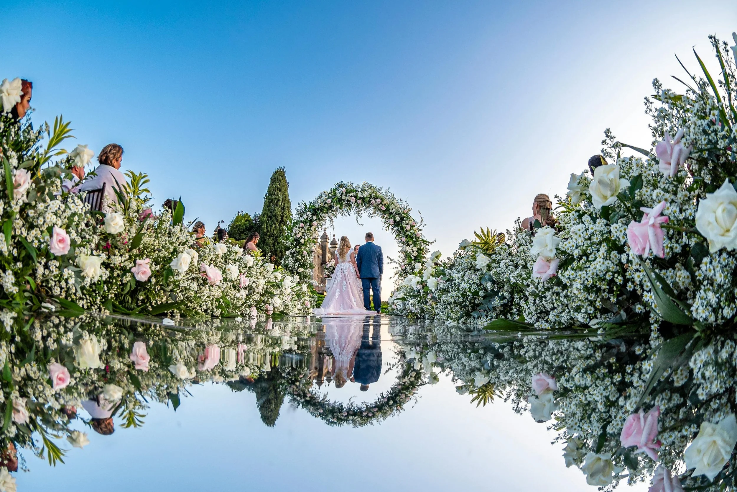 Bride and groom standing at an outdoor destination wedding ceremony under a floral arch, with abundant white and blush flowers lining a mirrored aisle at sunset