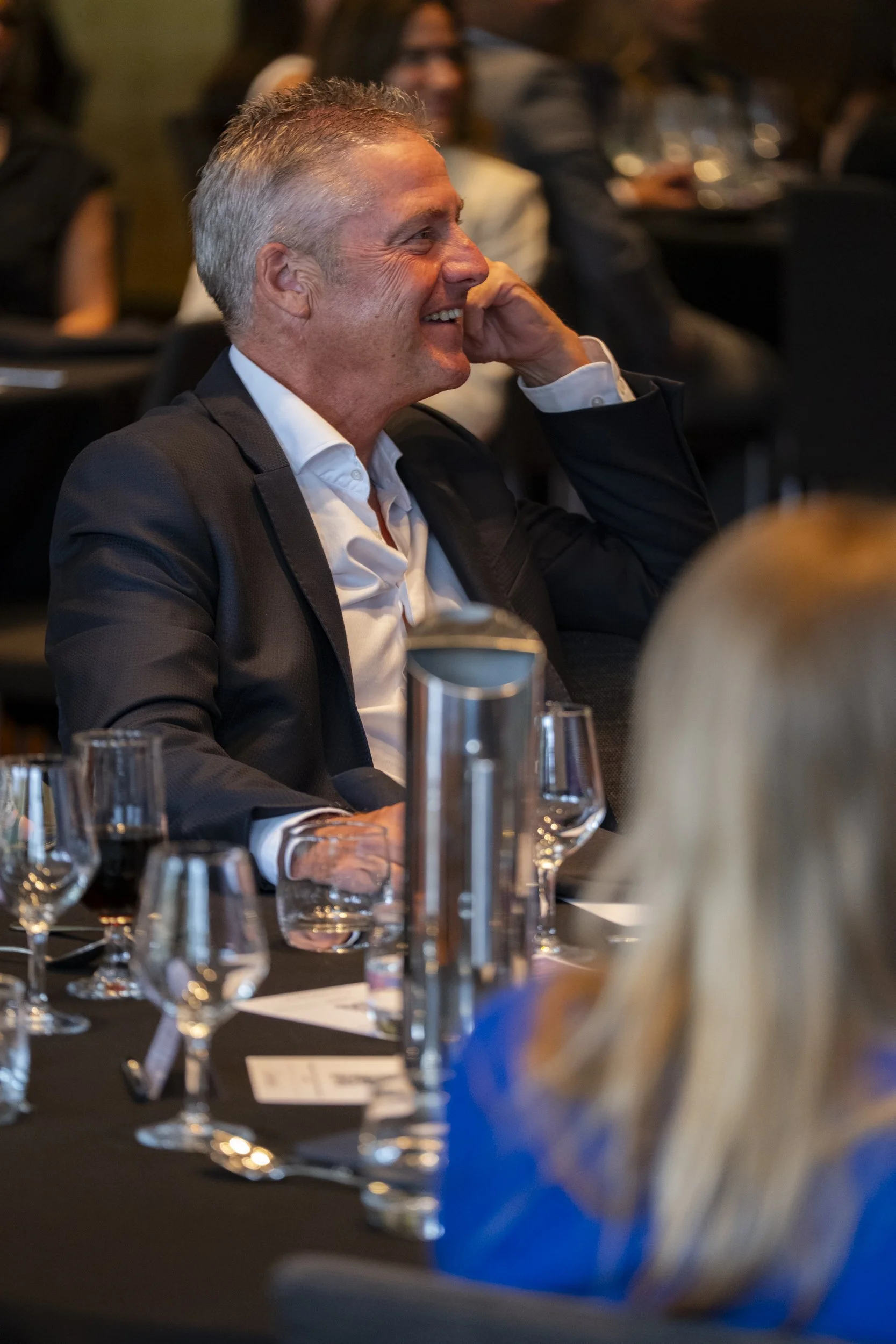 Male guest laughing at a corporate event dinner, seated at a formal table with glassware in the foreground