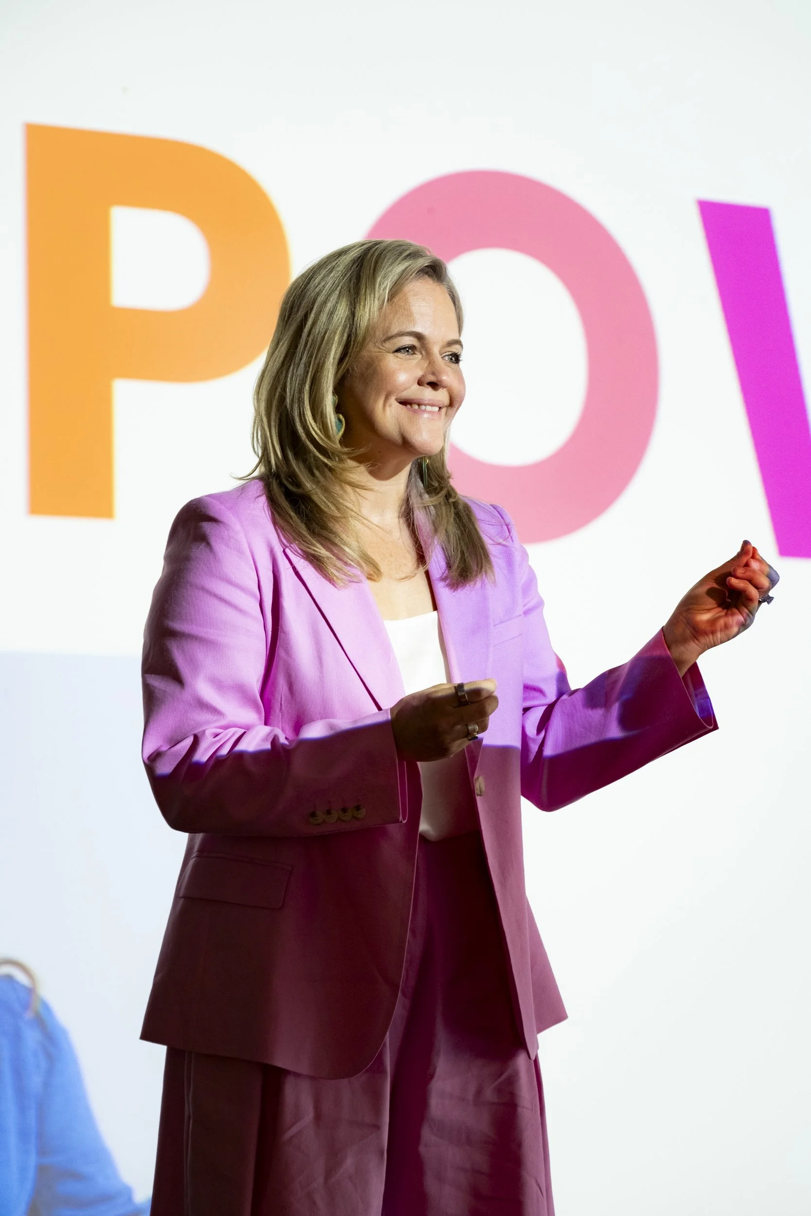 Close-up of a speaker mid-presentation on stage, smiling and gesturing, with colourful event branding projected behind her