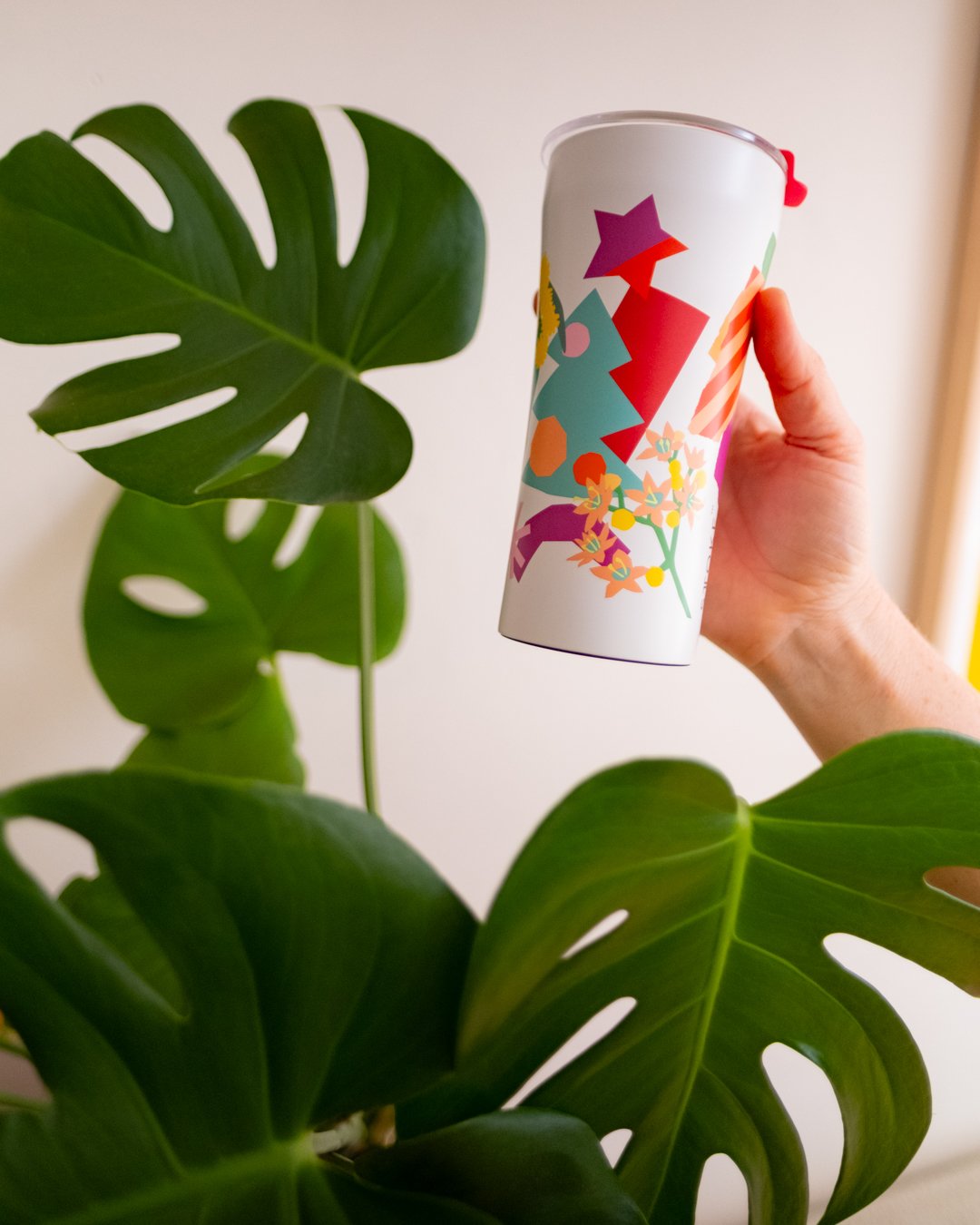 A hand holds a white STTOKE insulated tumbler with a bold collage-style print featuring geometric shapes and florals, held up against a large monstera plant