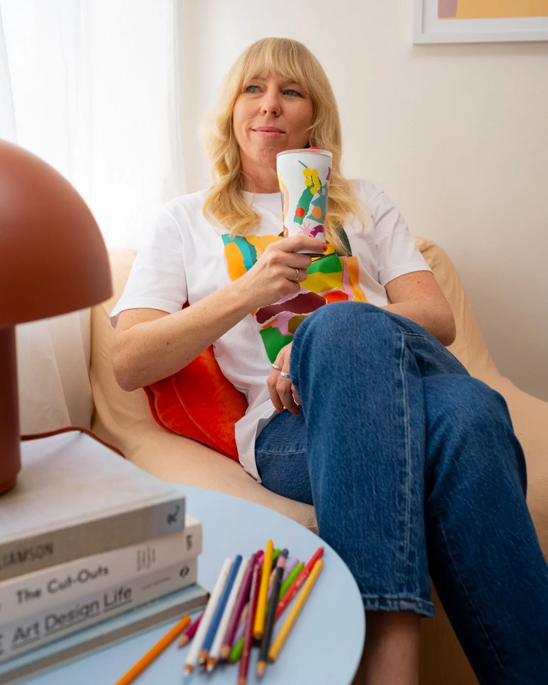 Leah Bartholomew sits in an armchair holding a STTOKE tumbler, with art books and coloured pencils on a coffee table in the foreground