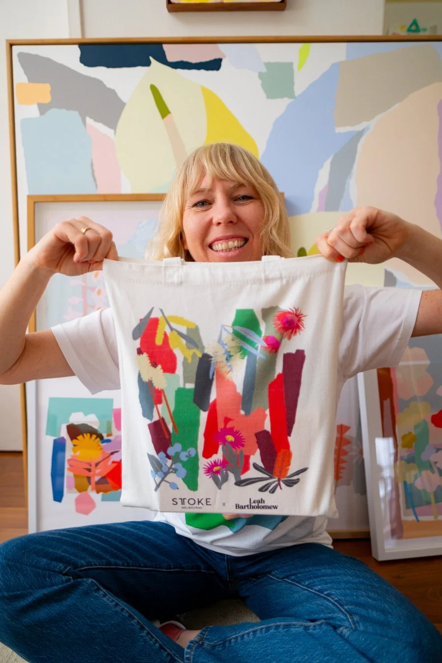 Leah Bartholomew sits on her studio floor, beaming as she holds up the STTOKE x Leah Bartholomew tote bag, with her large-scale abstract artworks visible behind her
