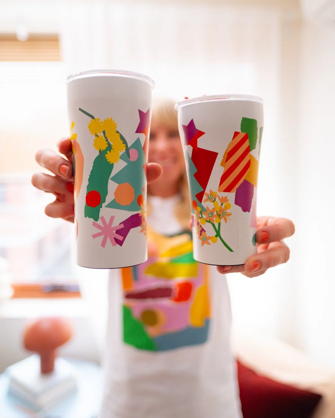 Leah Bartholomew holds up two white STTOKE tumblers towards the camera, both featuring different designs from the STTOKE x Leah Bartholomew collaboration