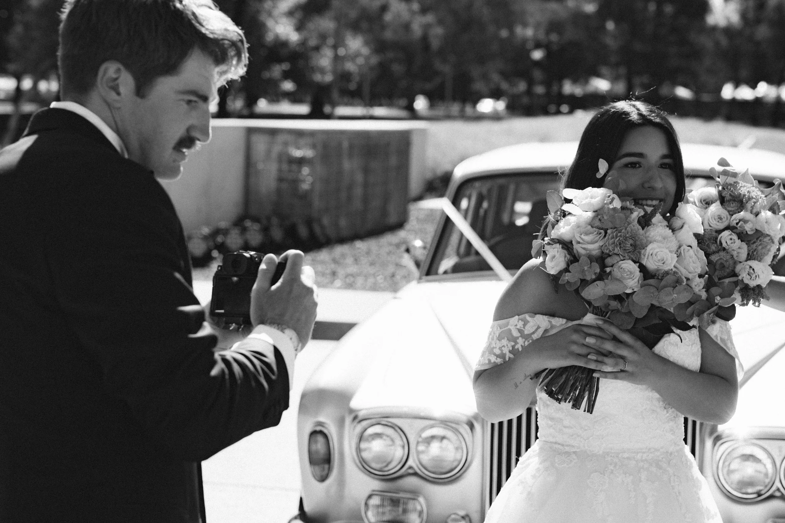 Black and white candid of groom photographing bride holding bouquet in front of a vintage Rolls-Royce