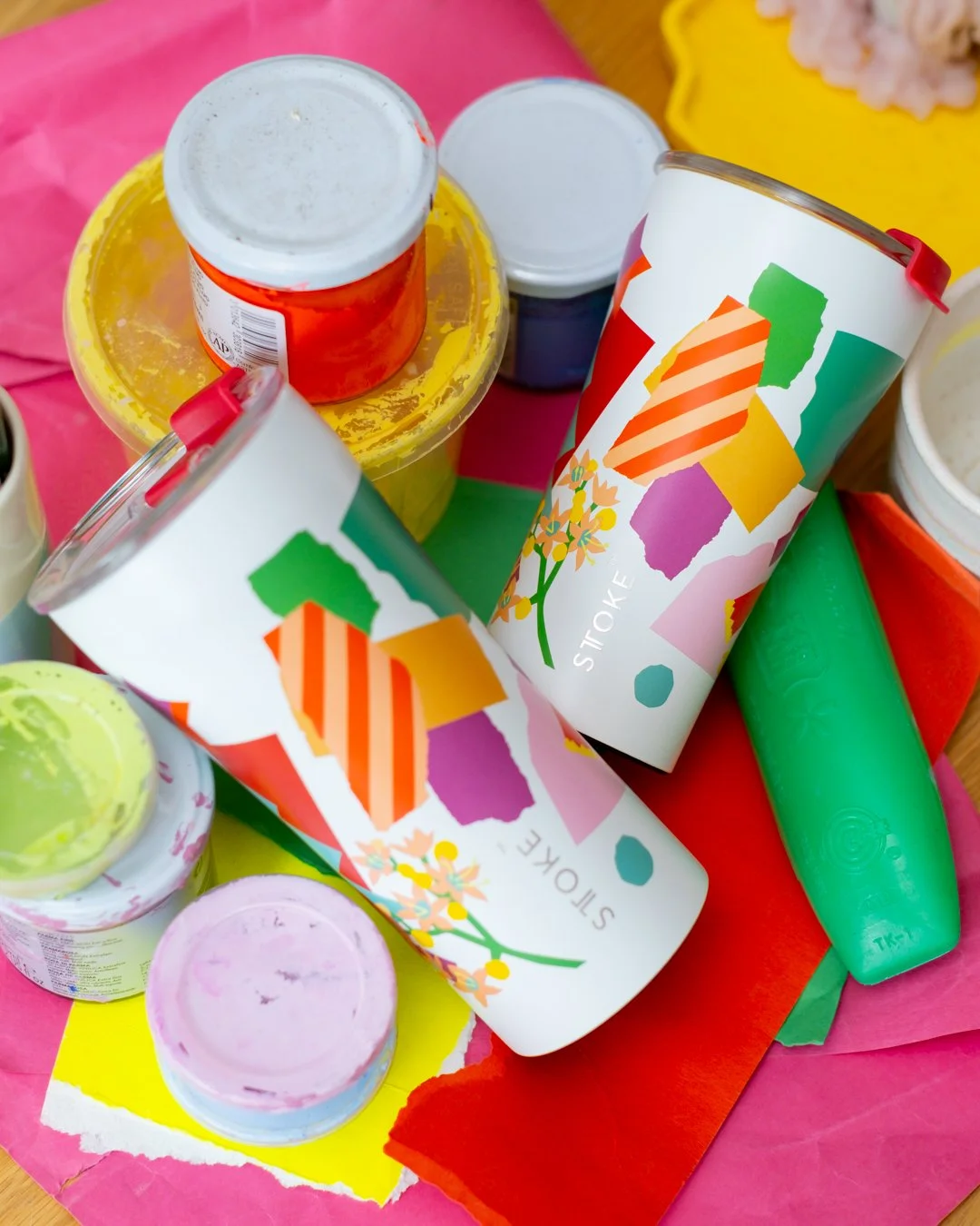 Two STTOKE tumblers featuring the Leah Bartholomew collaboration print laid among artist's paint pots, colourful torn paper scraps, and art supplies, shot from above on a bright pink background