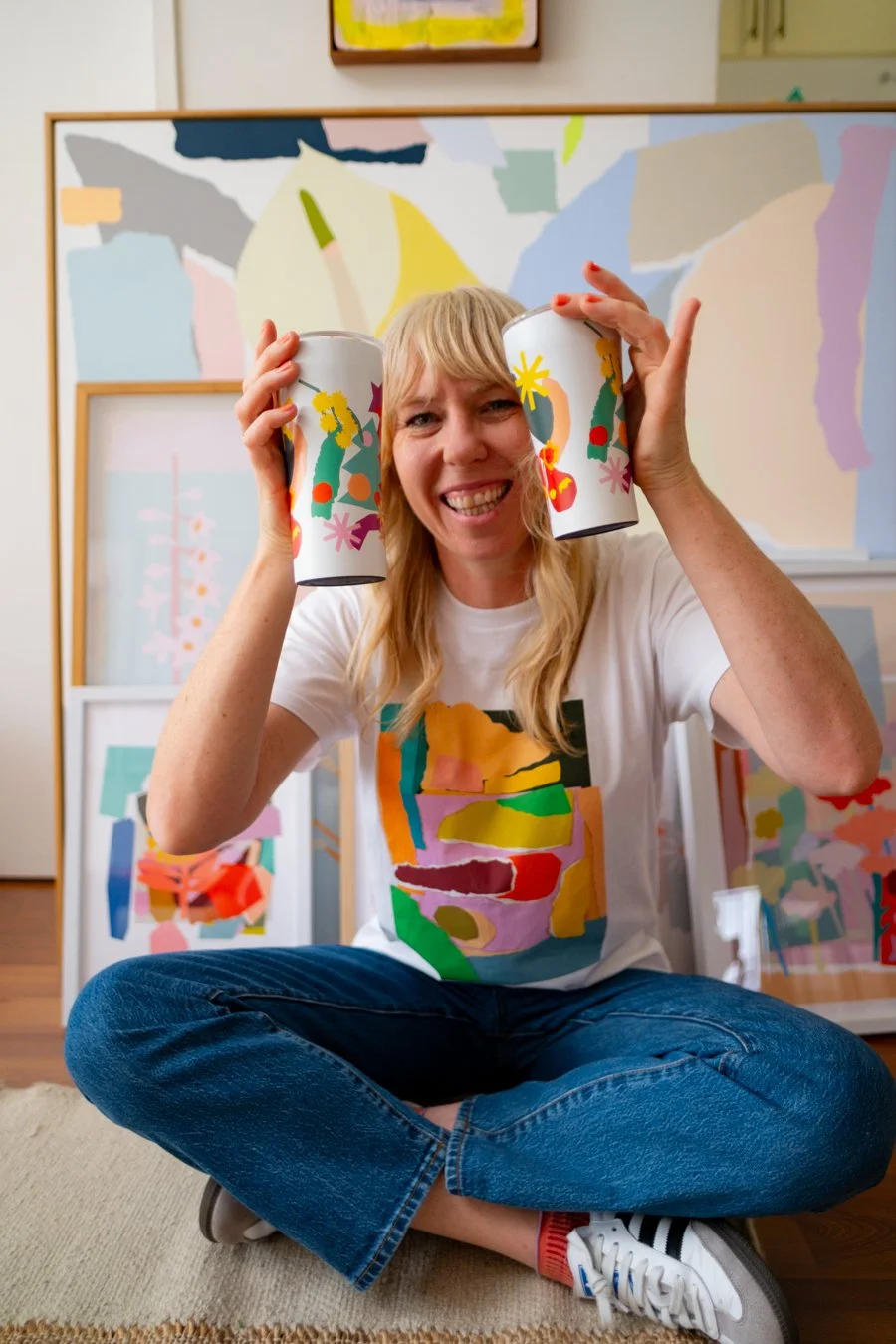 Leah Bartholomew sits cross-legged in her studio, holding two STTOKE tumblers up beside her face with a big smile, her abstract artworks displayed behind her