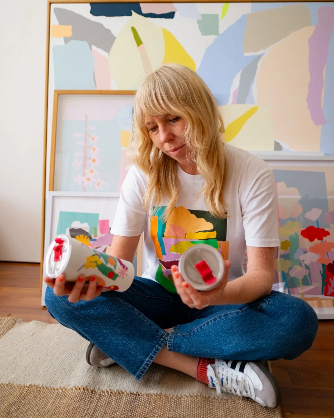 Leah Bartholomew sits cross-legged on the floor of her art studio, examining two STTOKE tumblers, surrounded by her framed abstract artworks