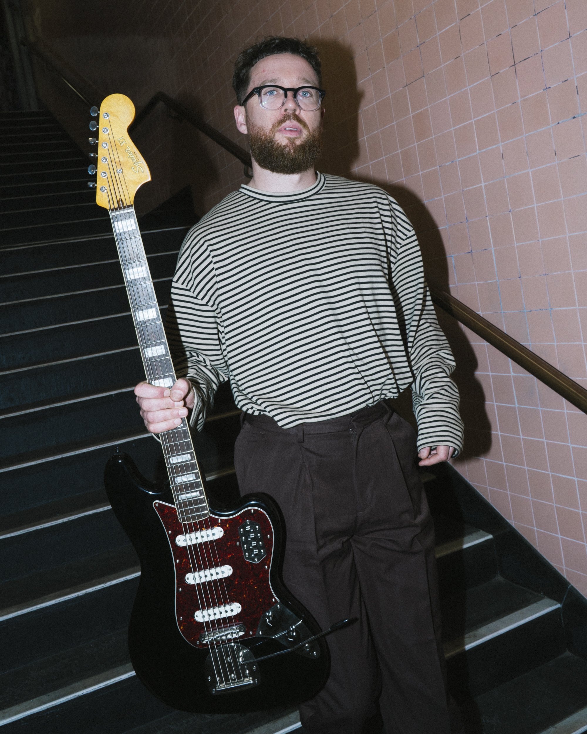 Musician holding a Fender Jaguar guitar beside a Telephones sign, pink tiled wall beneath Flinders Street Station