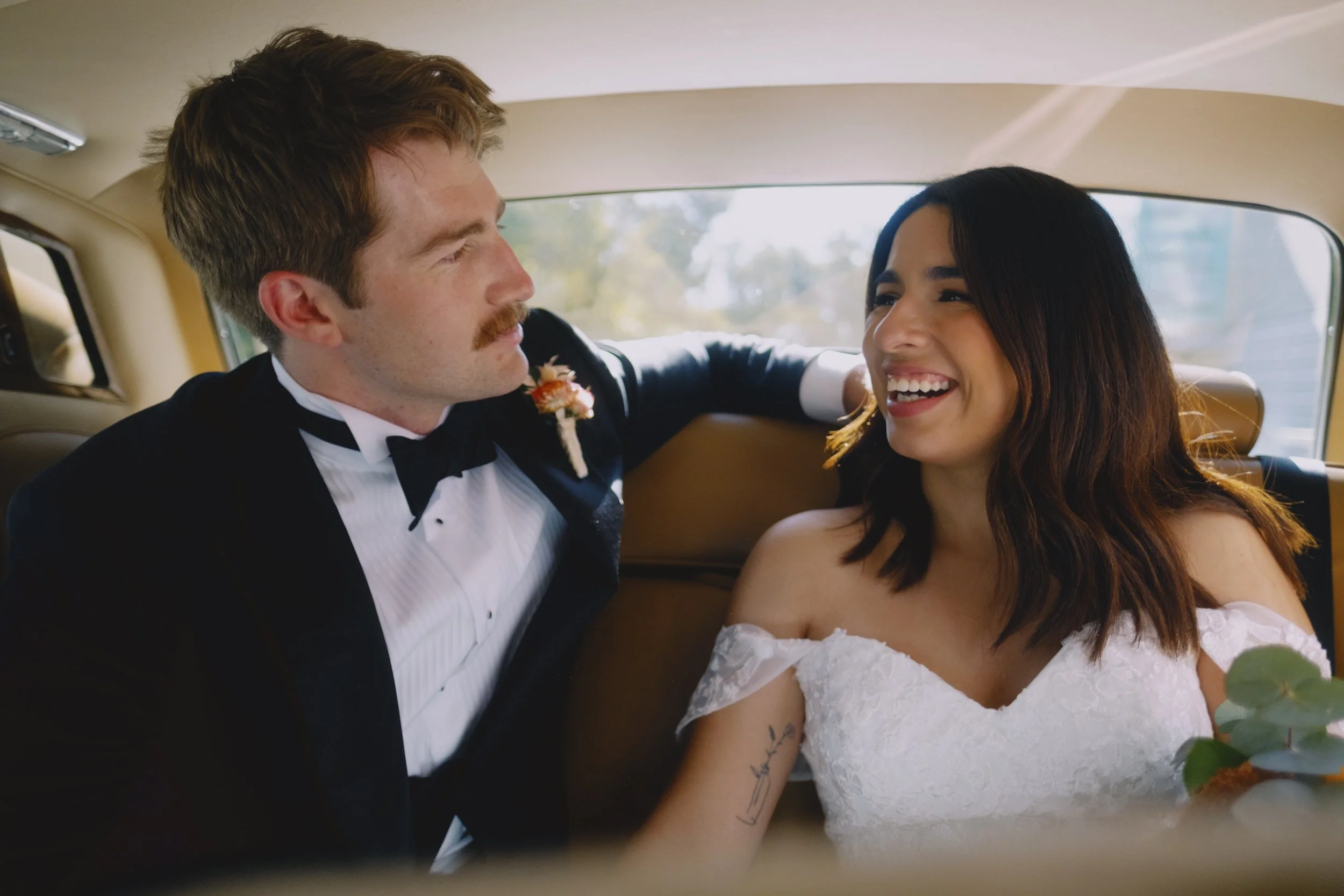 Bride and groom laughing together in the back seat of a vintage Rolls-Royce wedding car, warm leather interior