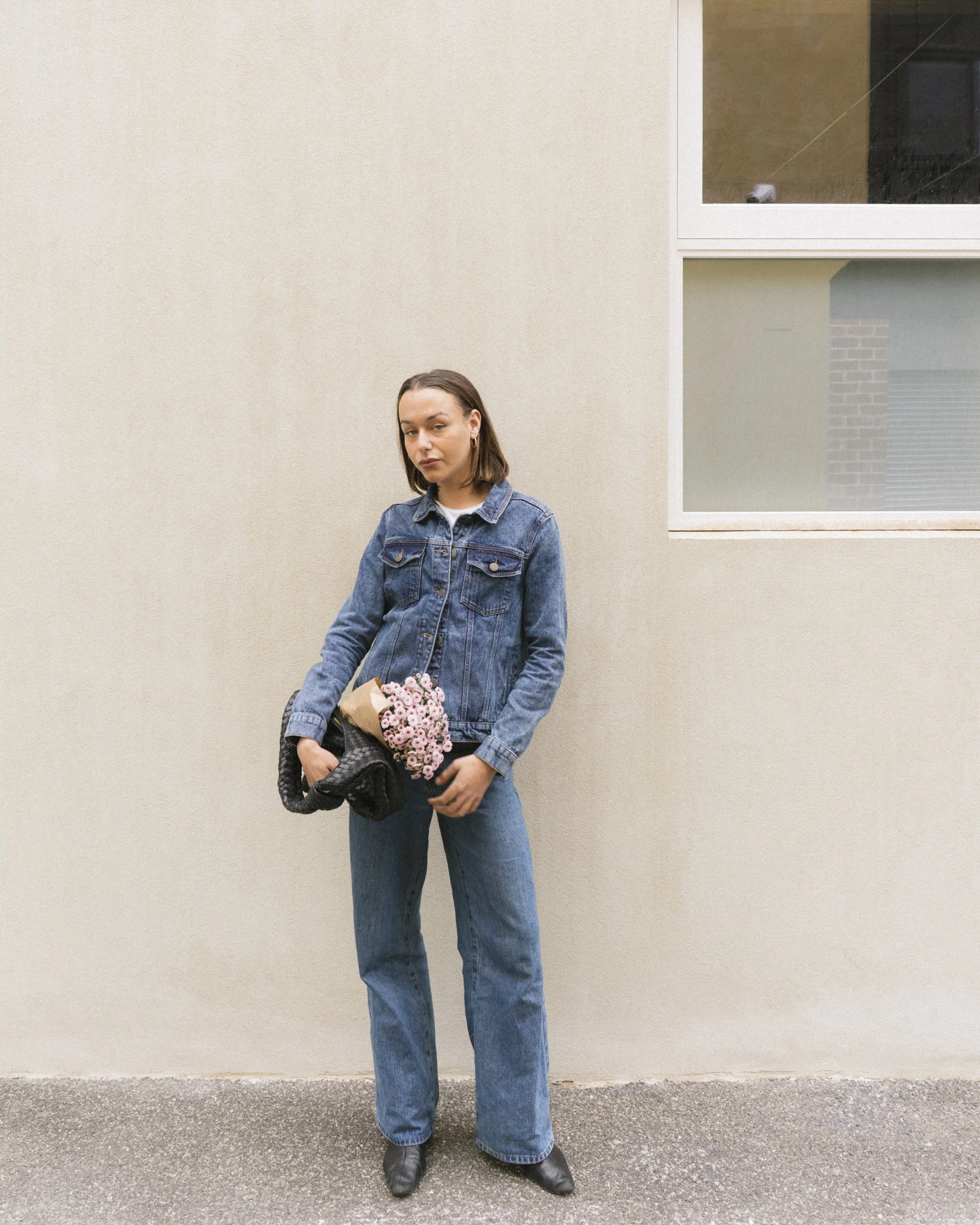 Full-length personal branding portrait of a woman in a denim jacket and jeans holding a wrapped bunch of pink flowers, leaning against a neutral render wall
