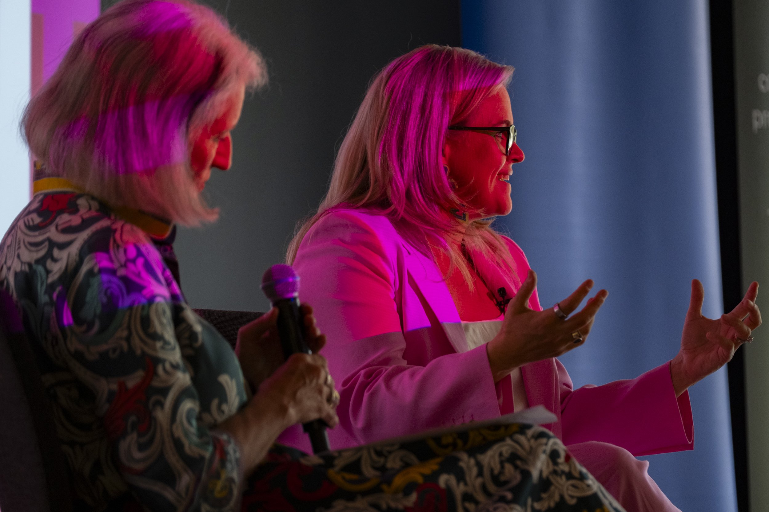 Two women on stage in conversation during a panel discussion, lit by vivid pink stage lighting at a corporate event