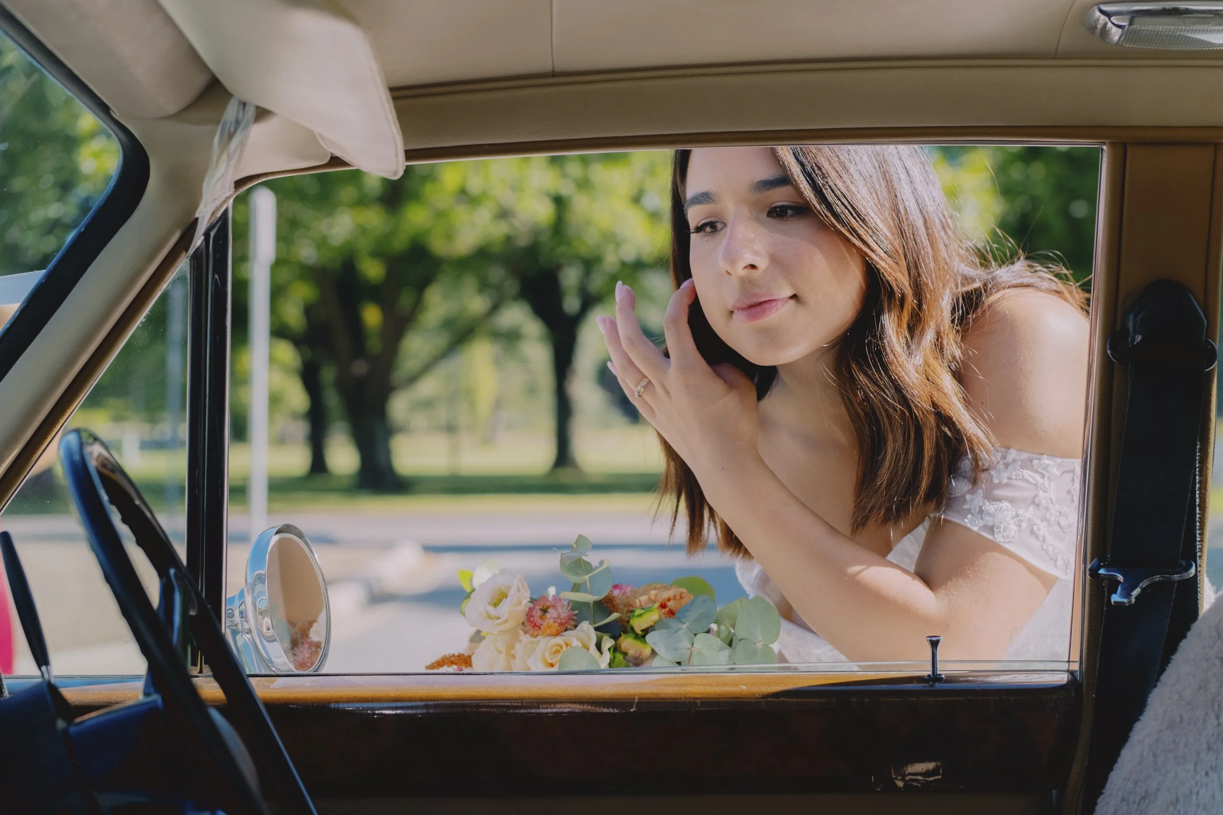 Bride leaning through the window of a vintage Rolls-Royce, bouquet resting on the door, trees visible in background