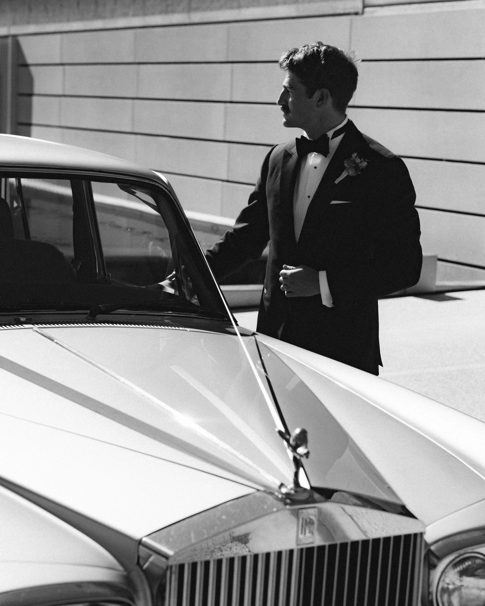 Black and white groom portrait beside a Rolls-Royce, opening the car door, strong directional sunlight