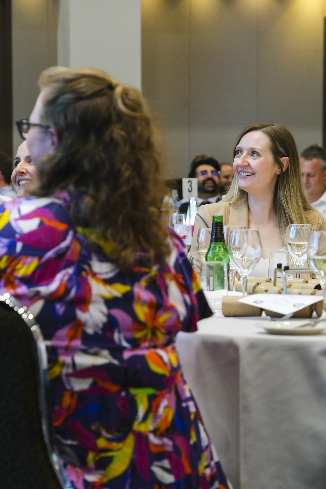 Corporate event guest smiling and watching a presentation at a formal luncheon, with other guests in the foreground