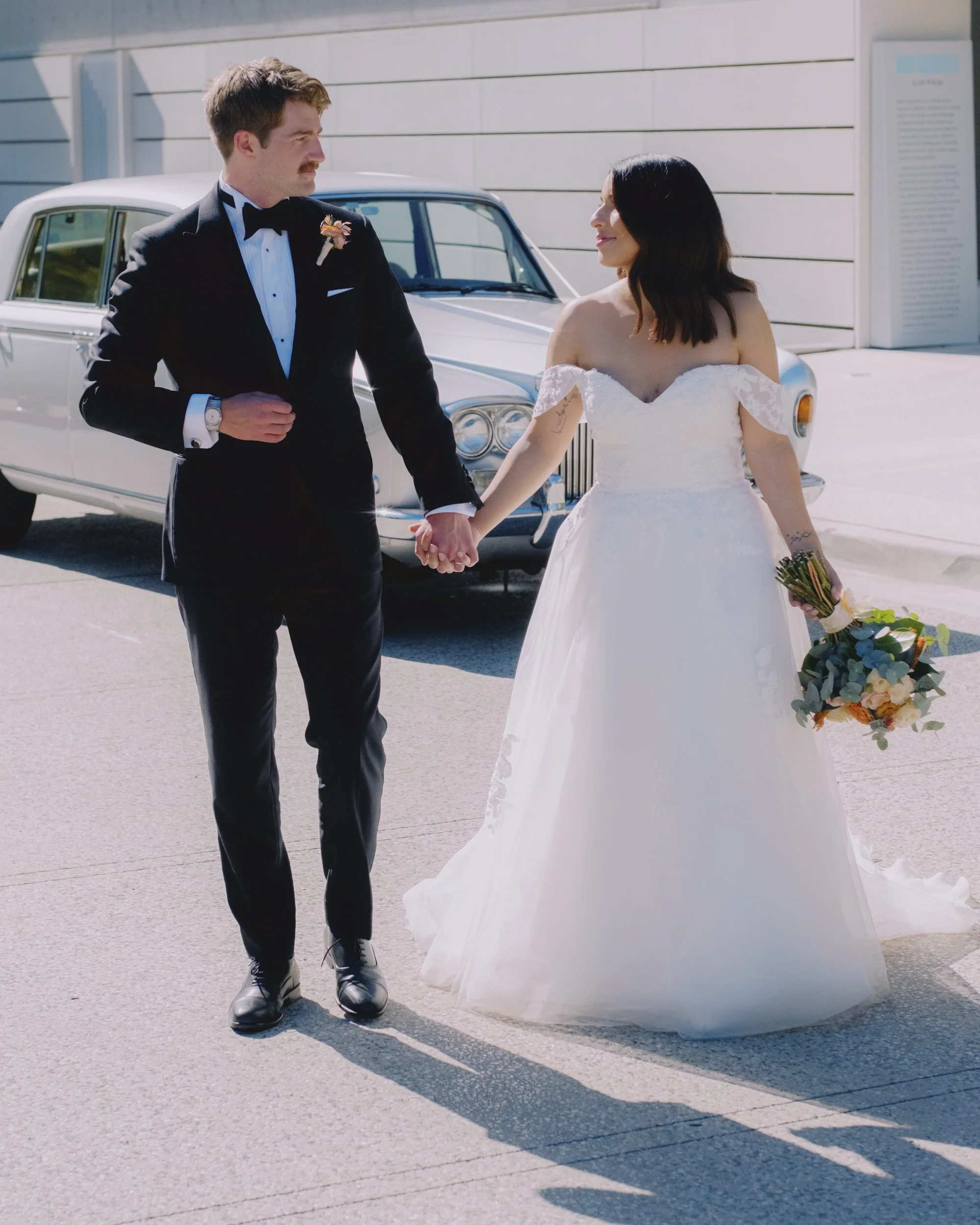 Bride and groom walking hand-in-hand beside a vintage Rolls-Royce, bride carrying an orange and peach bouquet, NGA precinct, Canberra