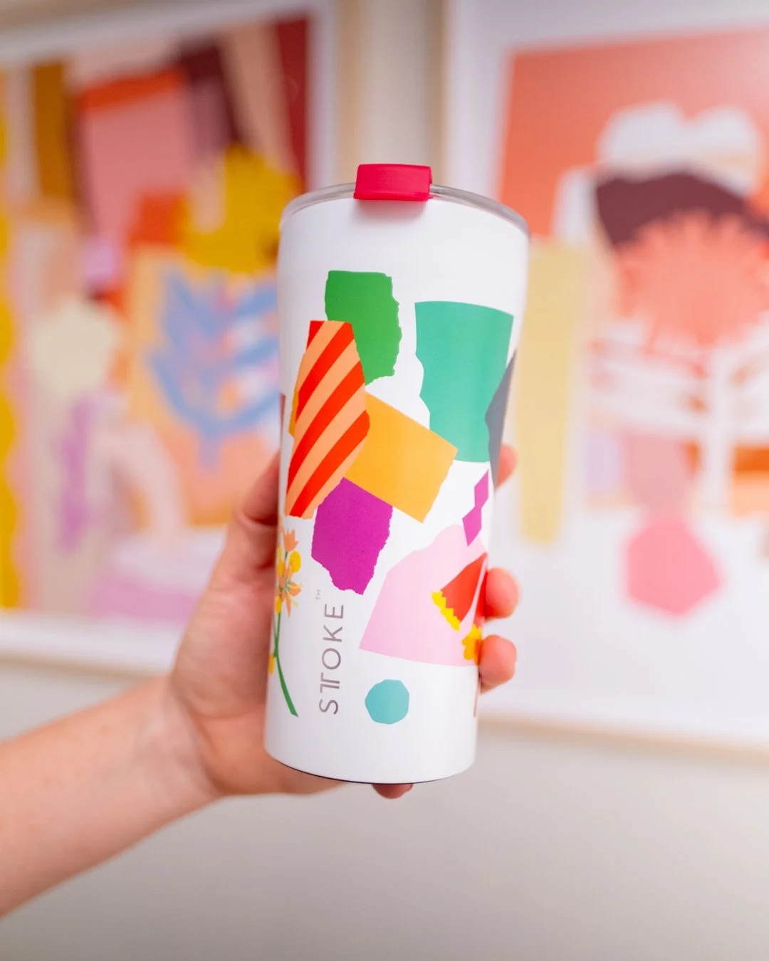 A hand holds a white STTOKE insulated tumbler featuring a vibrant collage-style print with colourful geometric shapes and florals, with abstract art prints blurred in the background