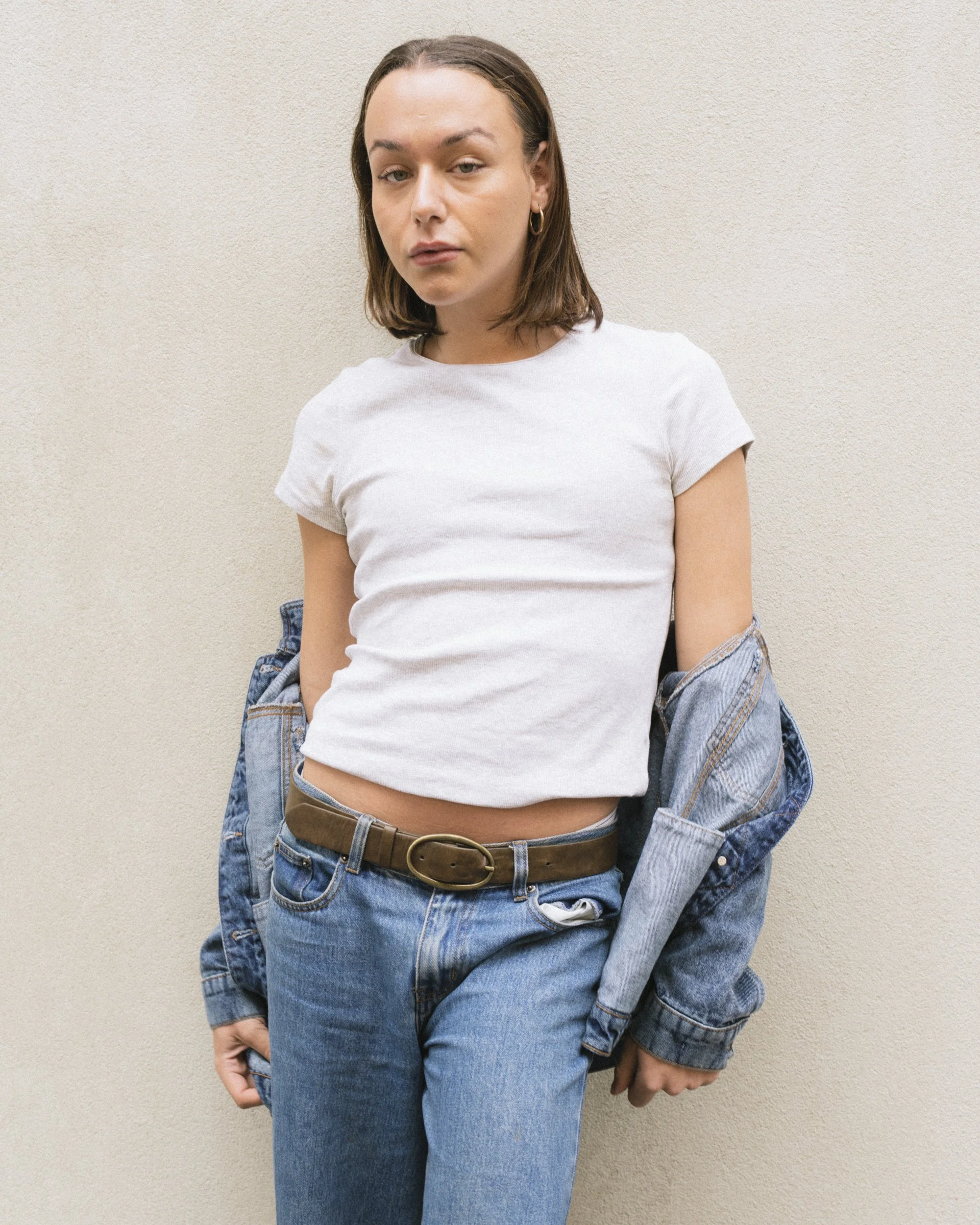Fashion portrait of a woman in a white t-shirt and low-rise jeans with a brown belt, denim jacket draped over her shoulder, against a neutral wall