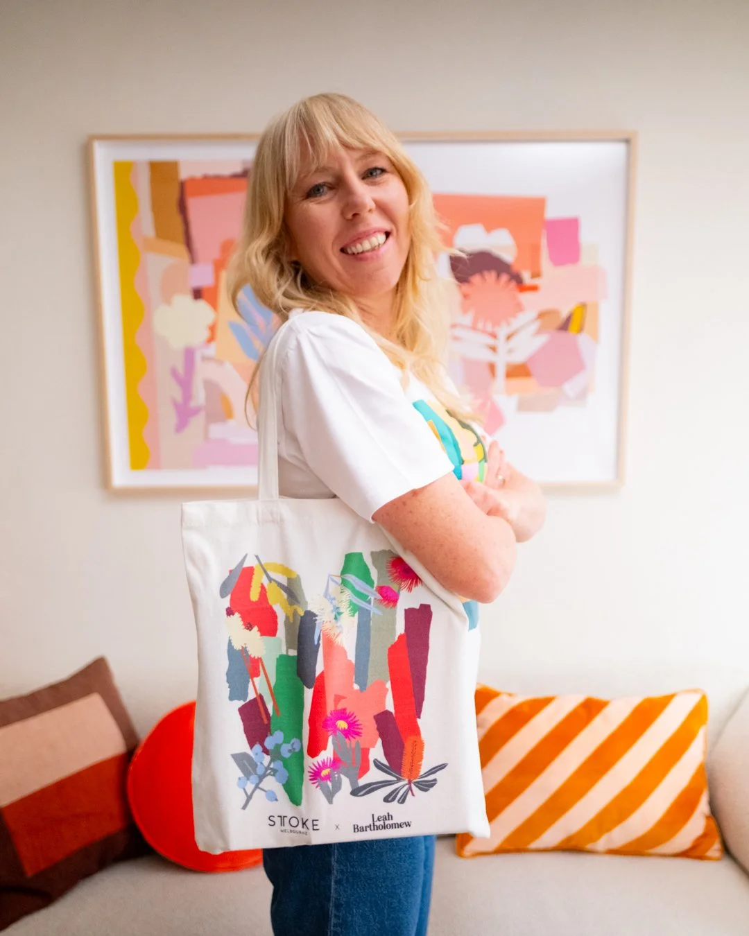 Leah Bartholomew smiles while wearing the STTOKE x Leah Bartholomew collaboration tote bag over her shoulder, standing in front of a large framed abstract artwork in her studio