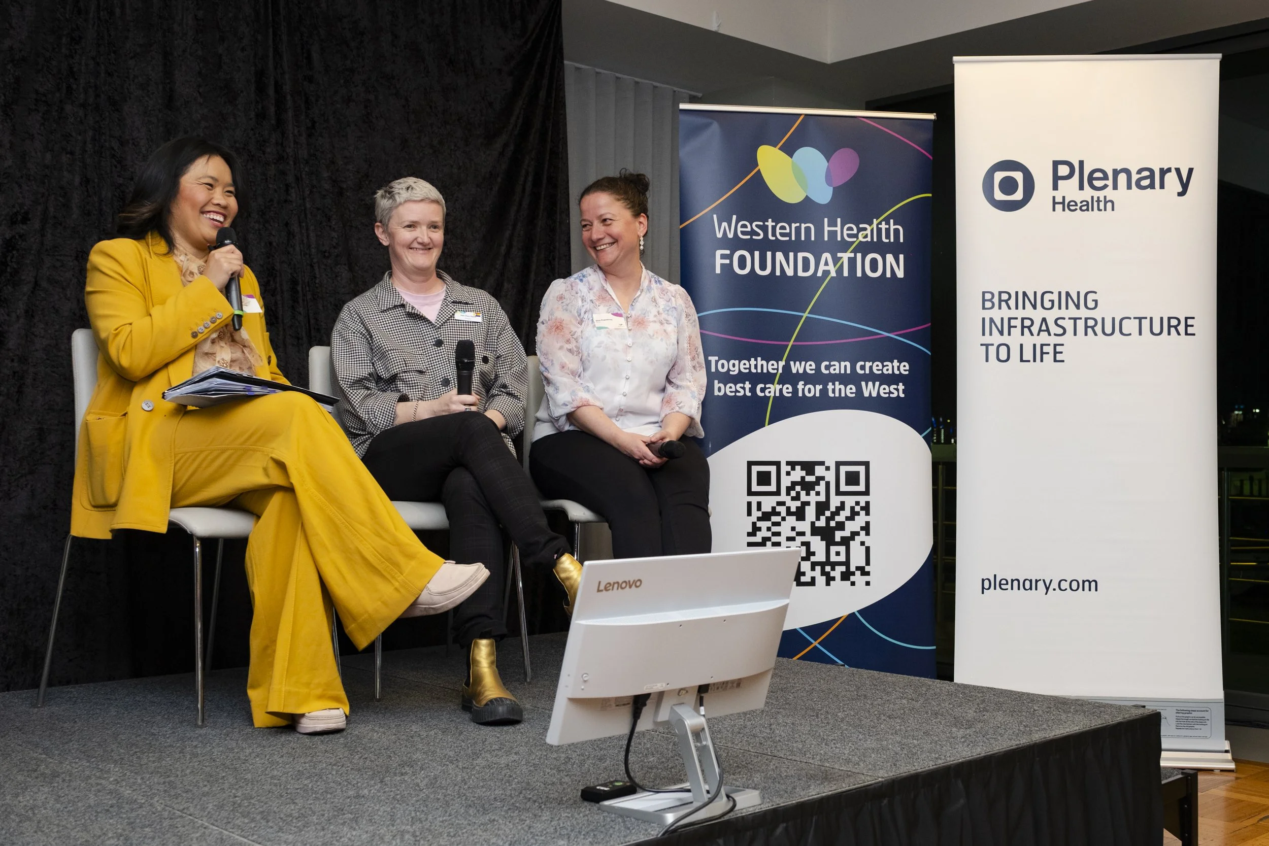 Three-person panel discussion on stage at a Western Health Foundation event, hosted by MC in yellow suit, with sponsor banners behind