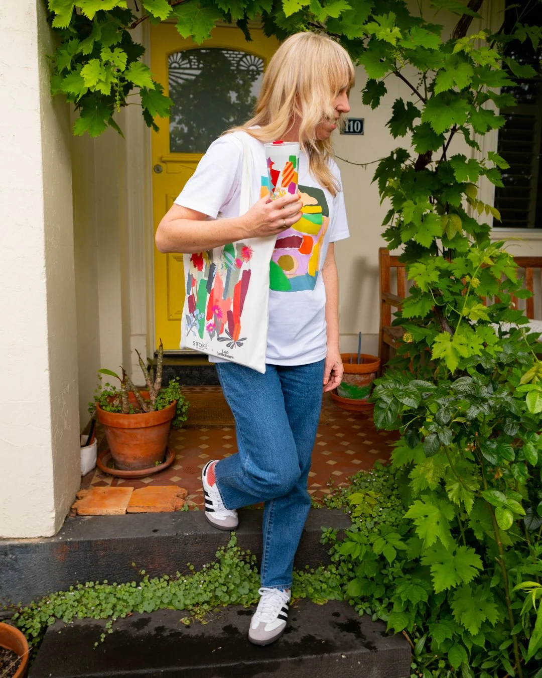 Leah Bartholomew steps out the front door of a Melbourne terrace house, holding a STTOKE tumbler and carrying the STTOKE x Leah Bartholomew tote bag, wearing jeans and Adidas sneakers