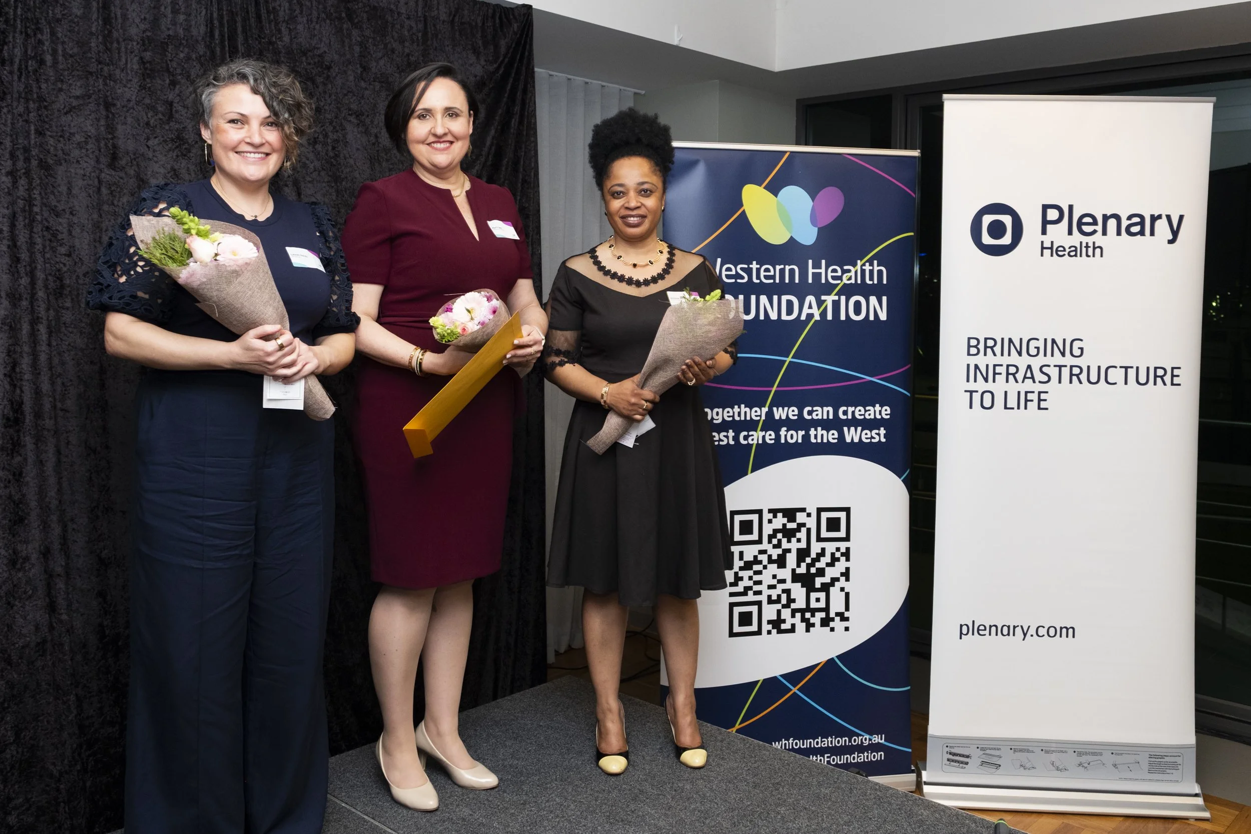 Three award recipients holding flowers and an award certificate at a Western Health Foundation event, with Plenary Health sponsor banner visible