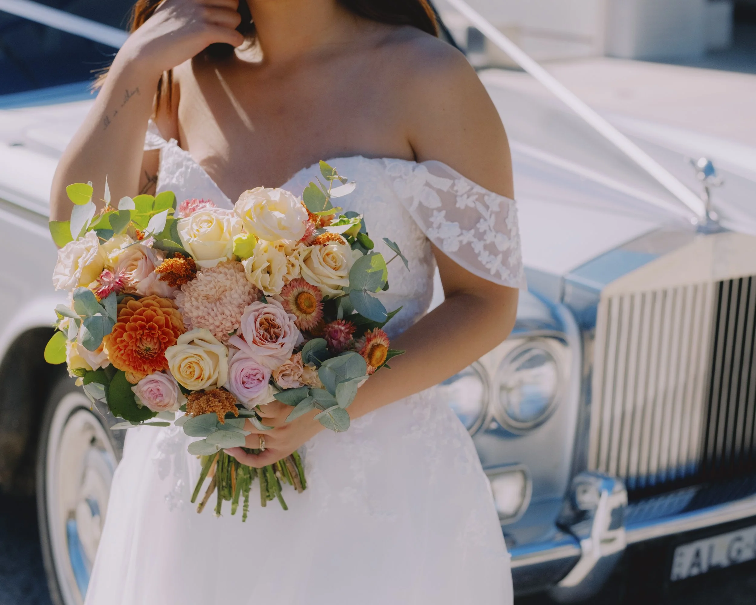 Close-up of bride holding a large orange, peach and cream wedding bouquet in front of a vintage Rolls-Royce