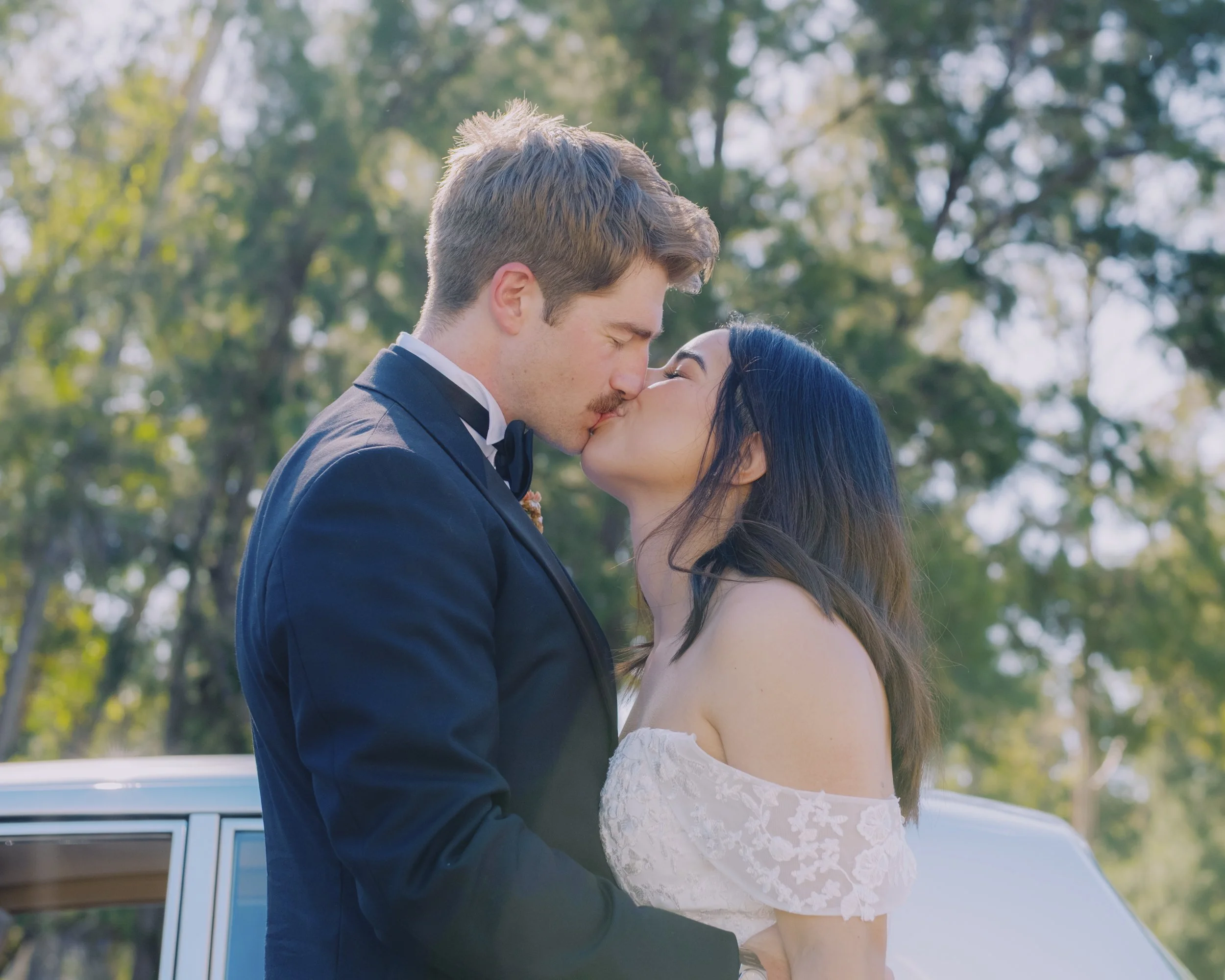 Bride and groom kissing beside a vintage Rolls-Royce, surrounded by trees in bright afternoon sunlight, Canberra