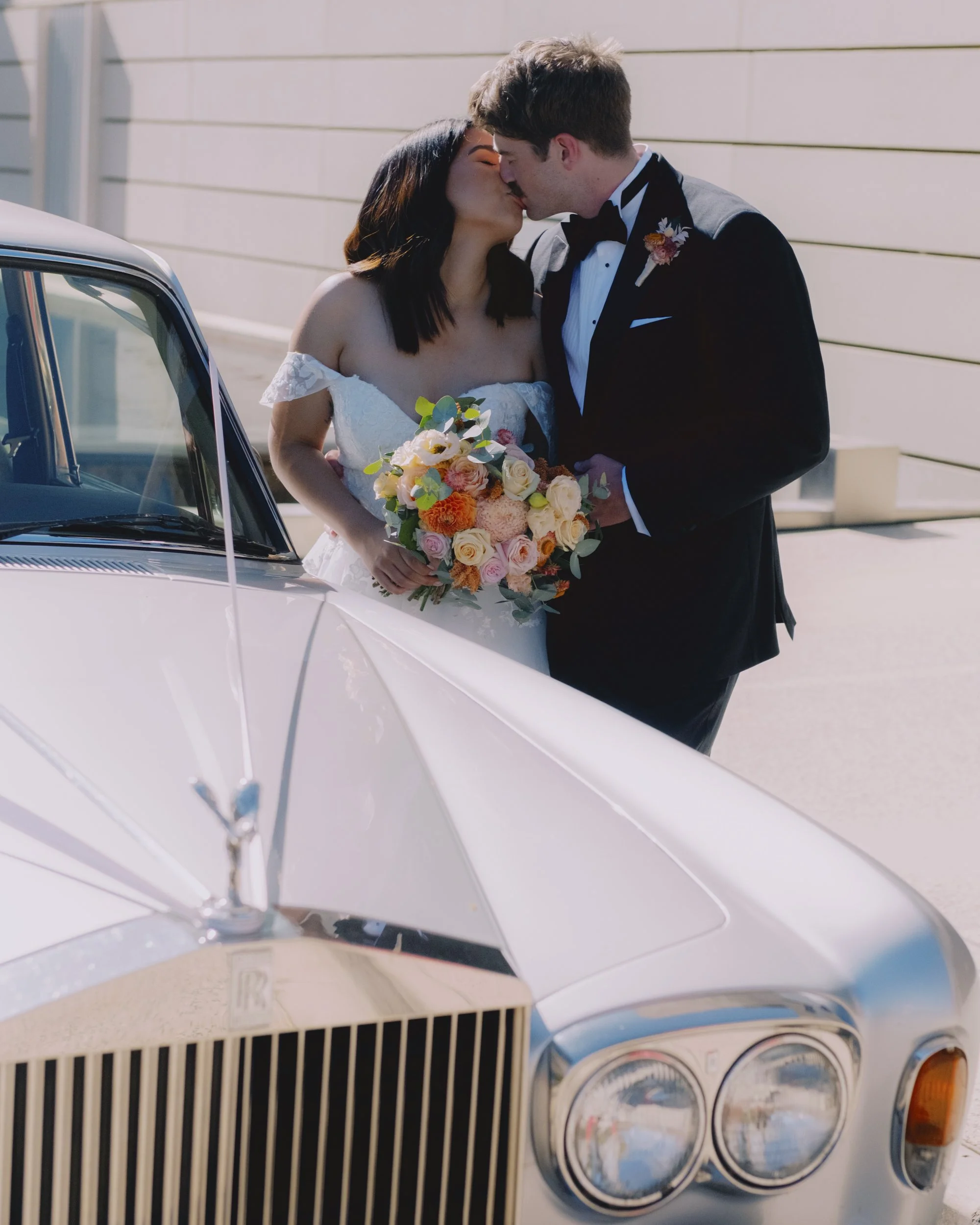 Bride and groom kissing over the bonnet of a vintage Rolls-Royce, bride holding orange and peach bouquet