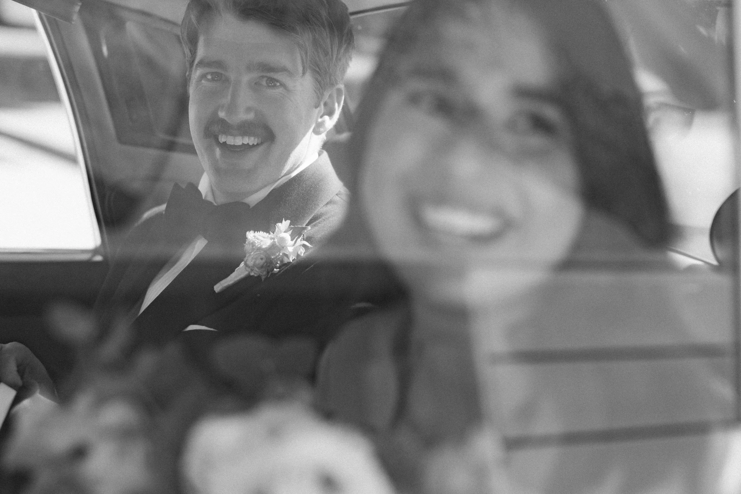 Black and white candid of bride and groom smiling together inside a vintage Rolls-Royce wedding car

