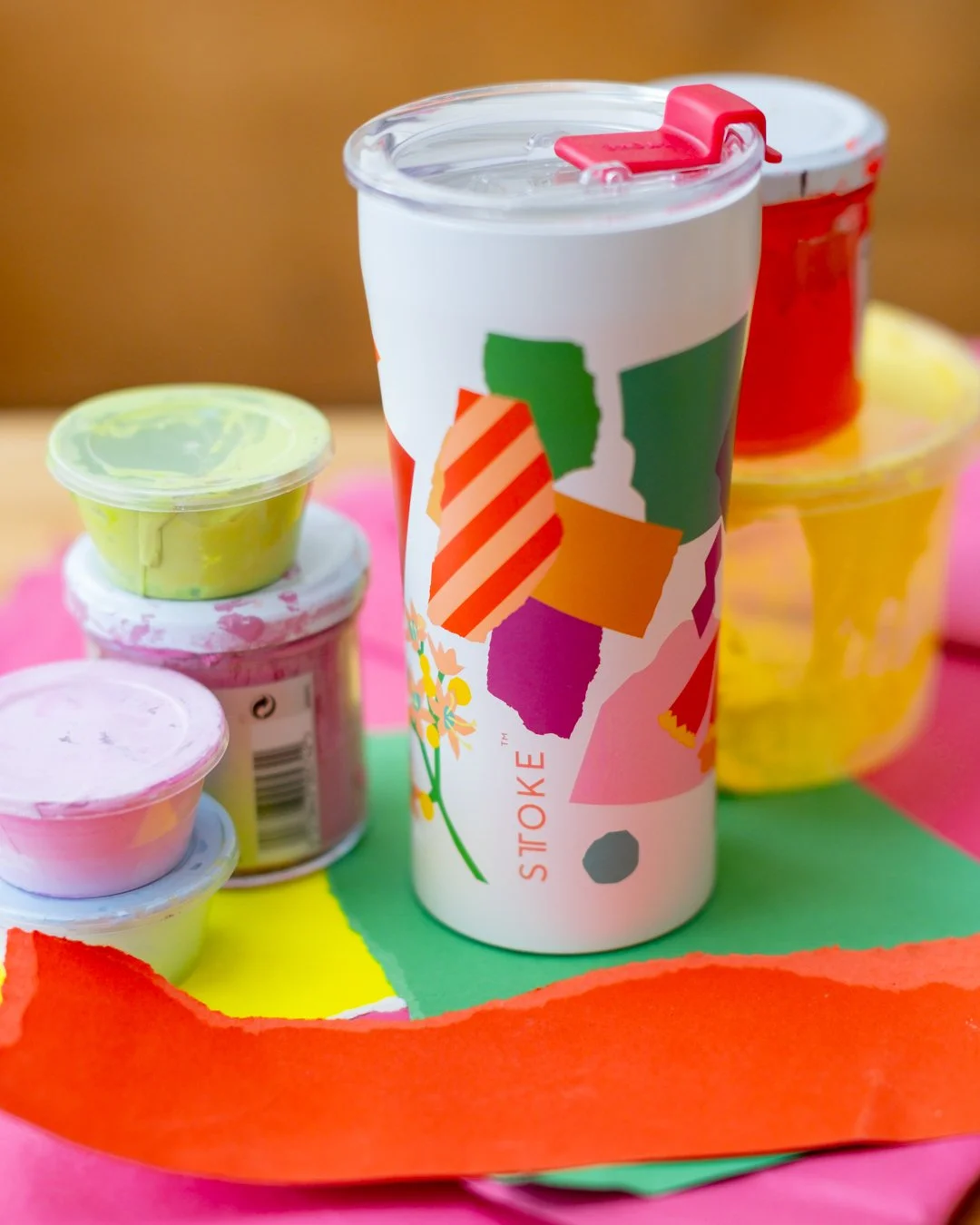 A white STTOKE tumbler with the Leah Bartholomew collaboration print — featuring colourful geometric shapes and florals — styled upright among stacked paint pots and torn coloured paper scraps on a vibrant studio surface
