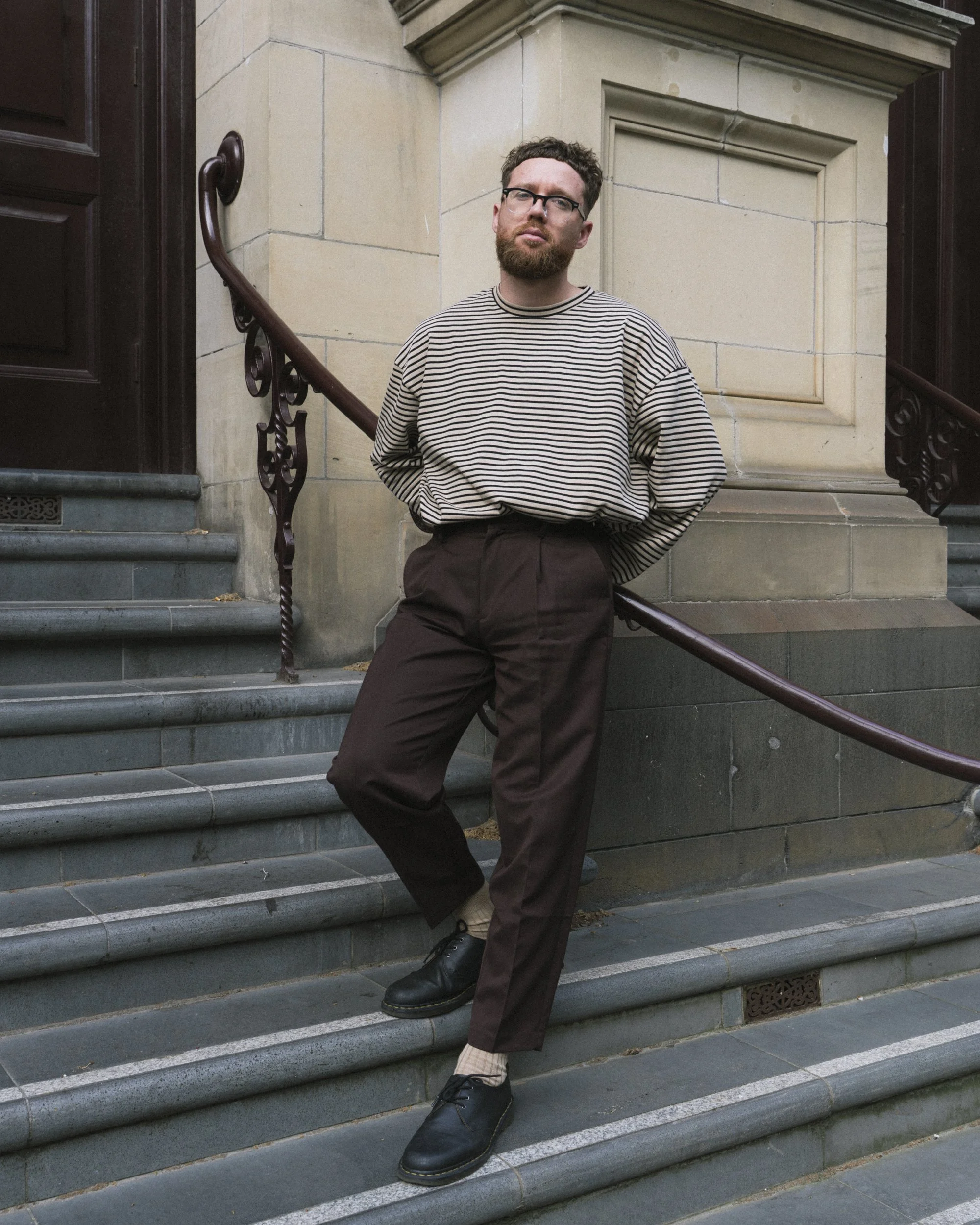 Full-length musician portrait on steps of heritage building, Melbourne CBD, striped jumper and brown trousers