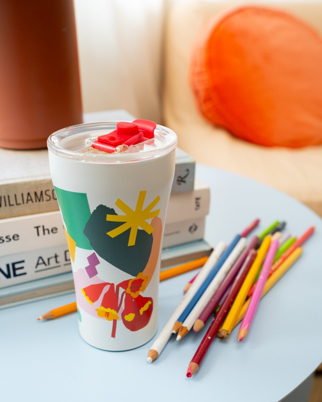 A white STTOKE tumbler featuring a bold collage-style print from the Leah Bartholomew collaboration, styled on a blue coffee table alongside coloured pencils and stacked art books