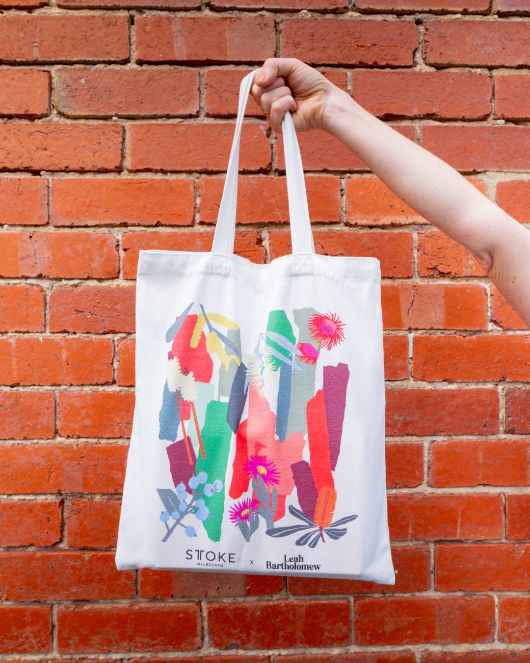 A hand holds up a STTOKE x Leah Bartholomew canvas tote bag against a red brick wall, showcasing the bold Australian native floral and abstract brushstroke print