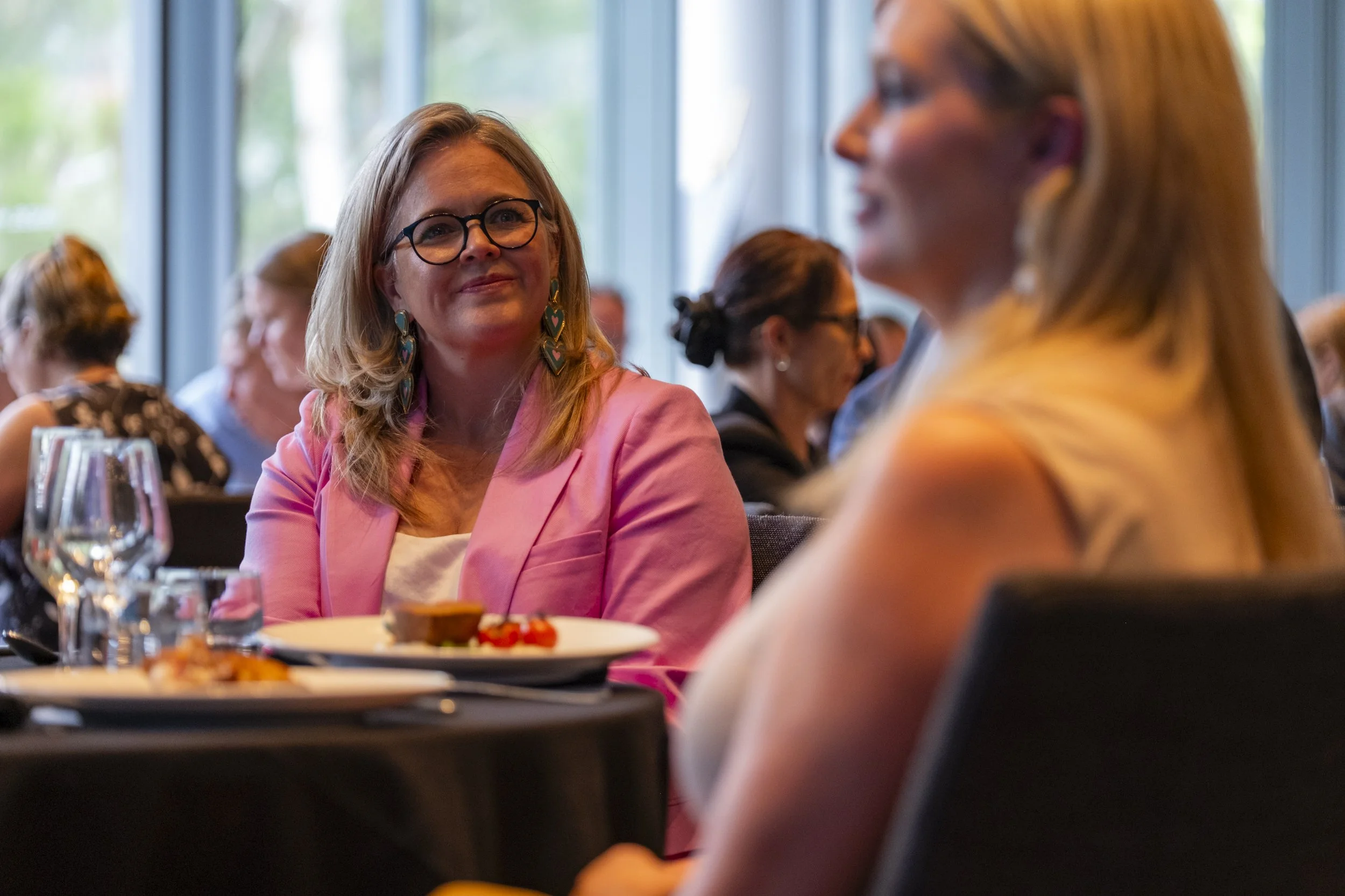 Event guest in a pink blazer listening attentively at a corporate luncheon, with a blurred foreground and window light behind