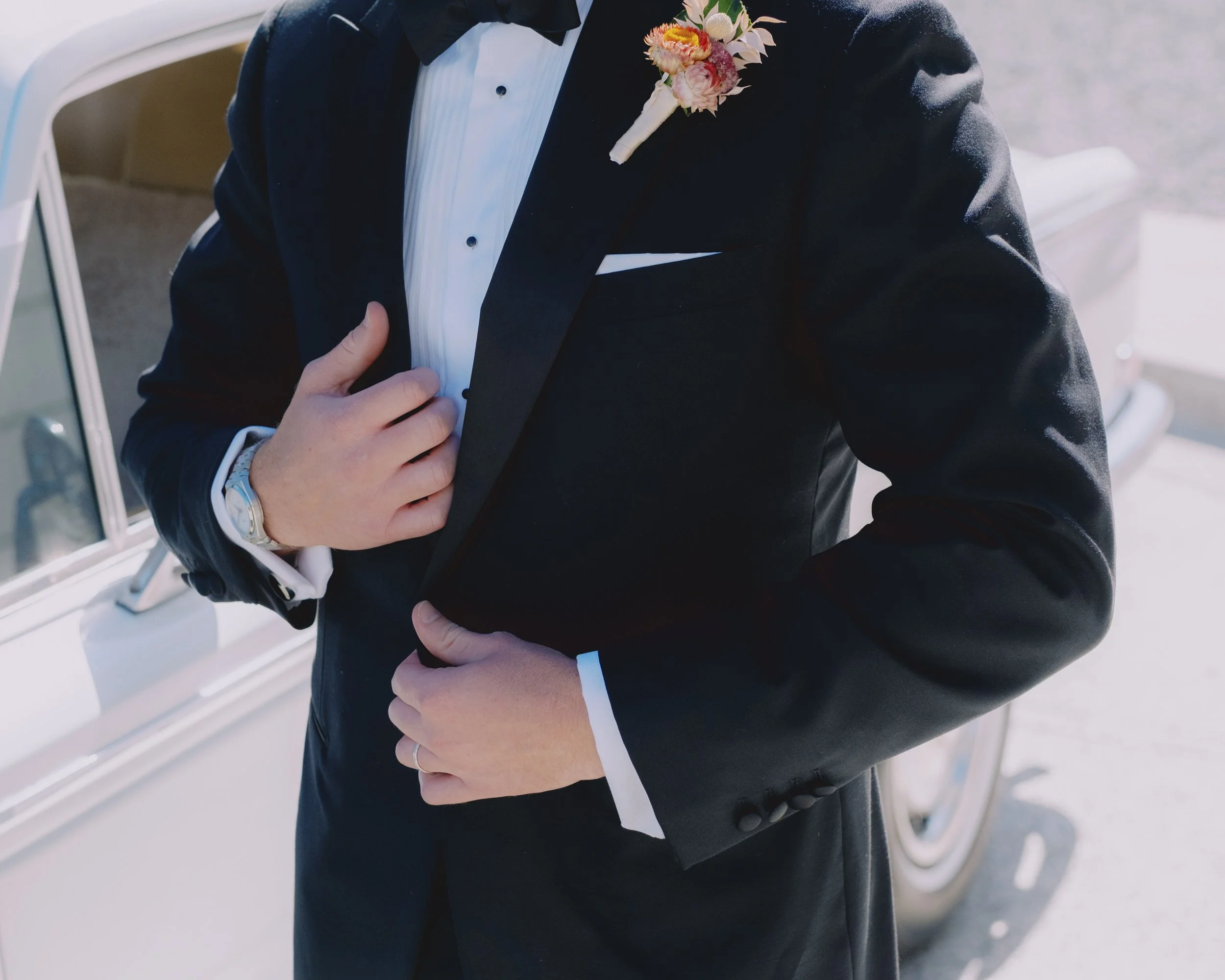 Detail shot of groom's tuxedo with floral boutonnière and watch, beside a vintage wedding car