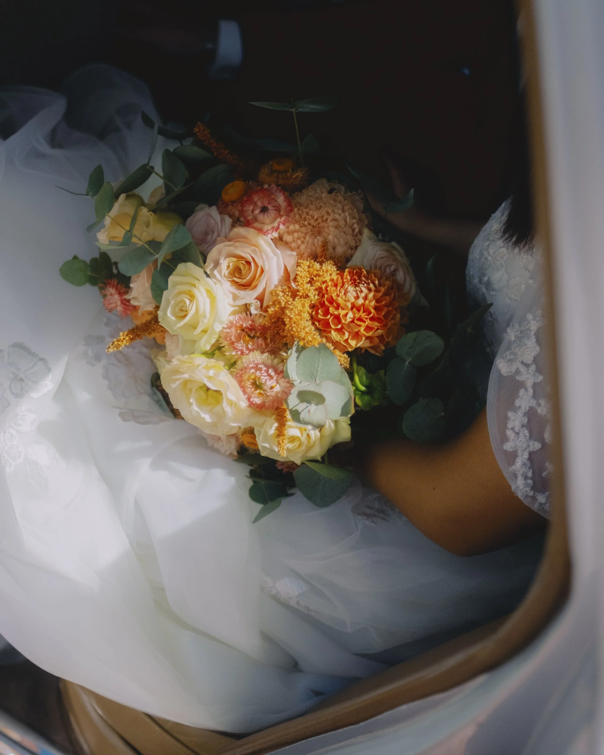 Wedding bouquet of orange dahlias, cream roses and eucalyptus resting on the bride's lap inside a vintage Rolls-Royce